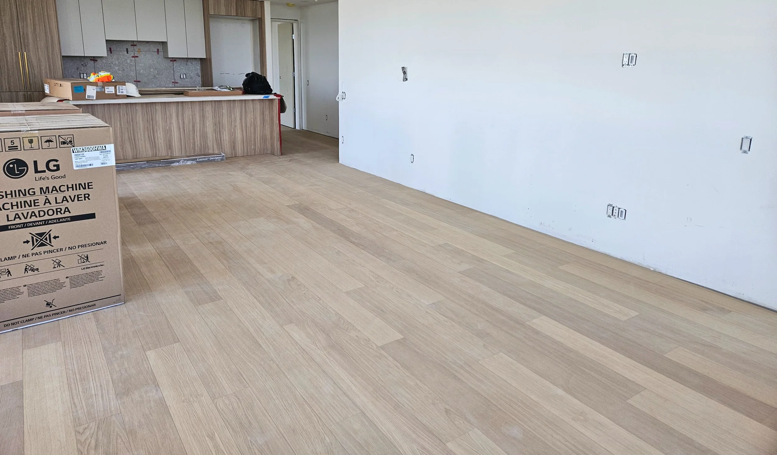 An unfinished kitchen with light wood flooring, a box for an LG washing machine, and some exposed electrical outlets on the wall.