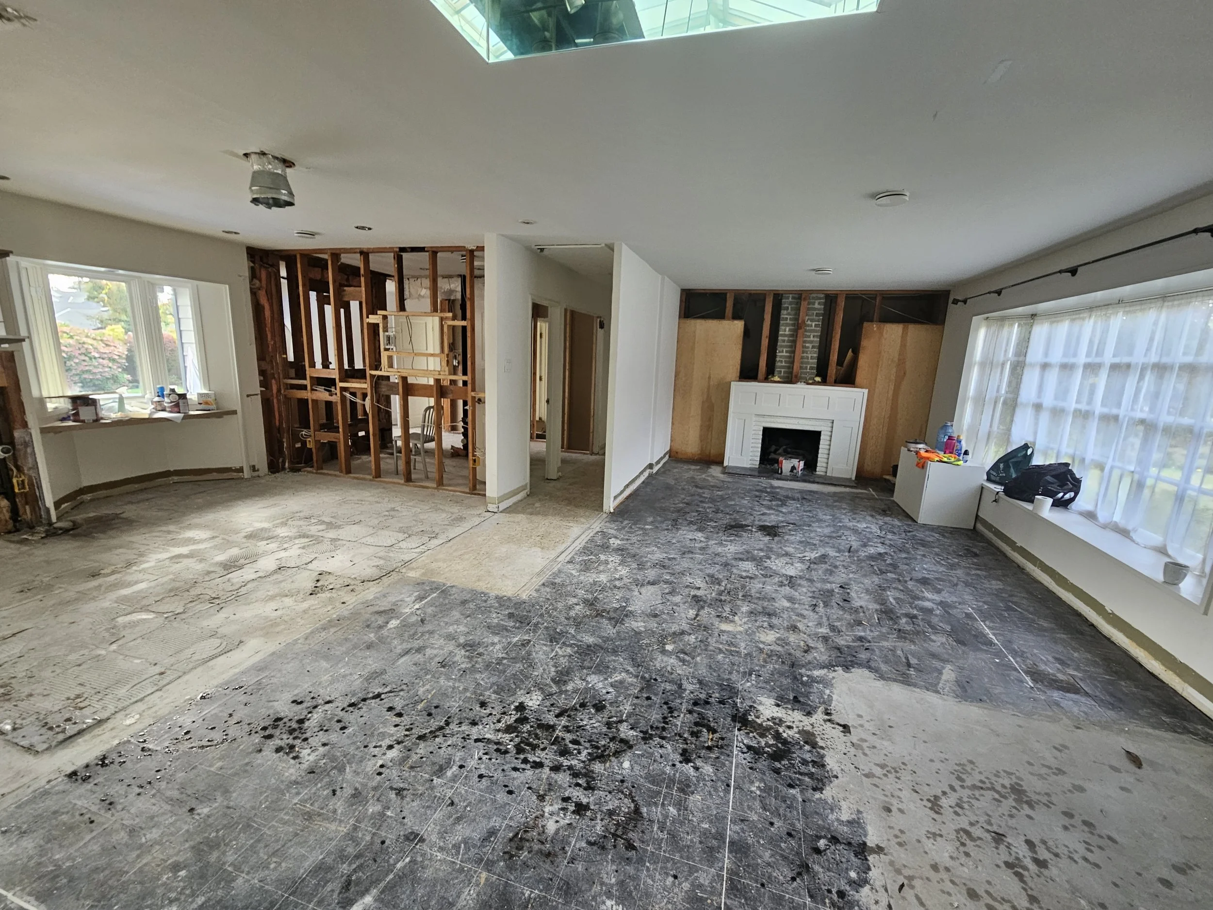 Living room under renovation with partially removed flooring, exposed wall framing, and a fireplace, with construction materials and tools on windowsills.