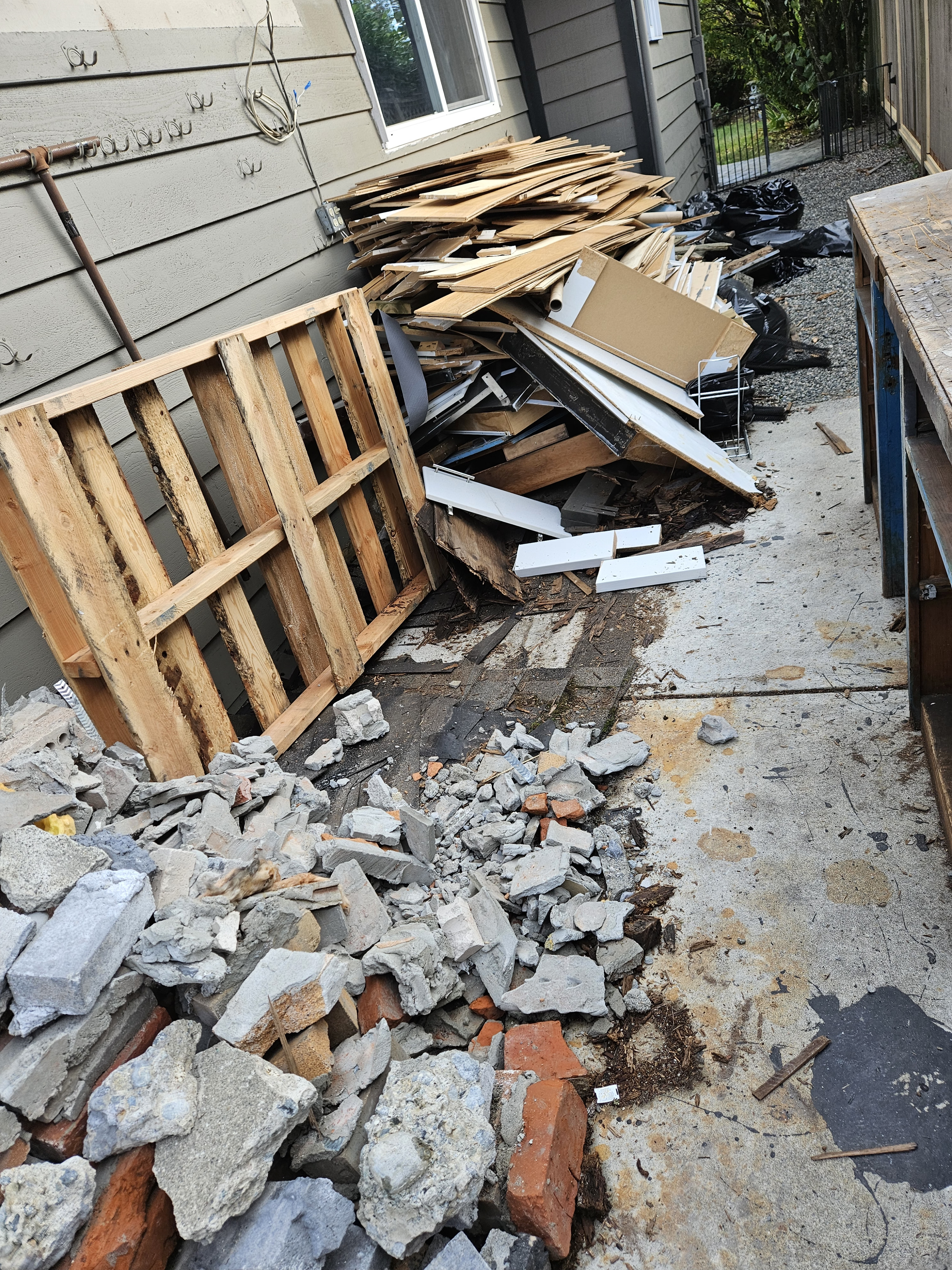 Piles of debris including rocks, concrete, wood, and cardboard in a backyard with a wooden pallet, black trash bags, and a workbench visible.
