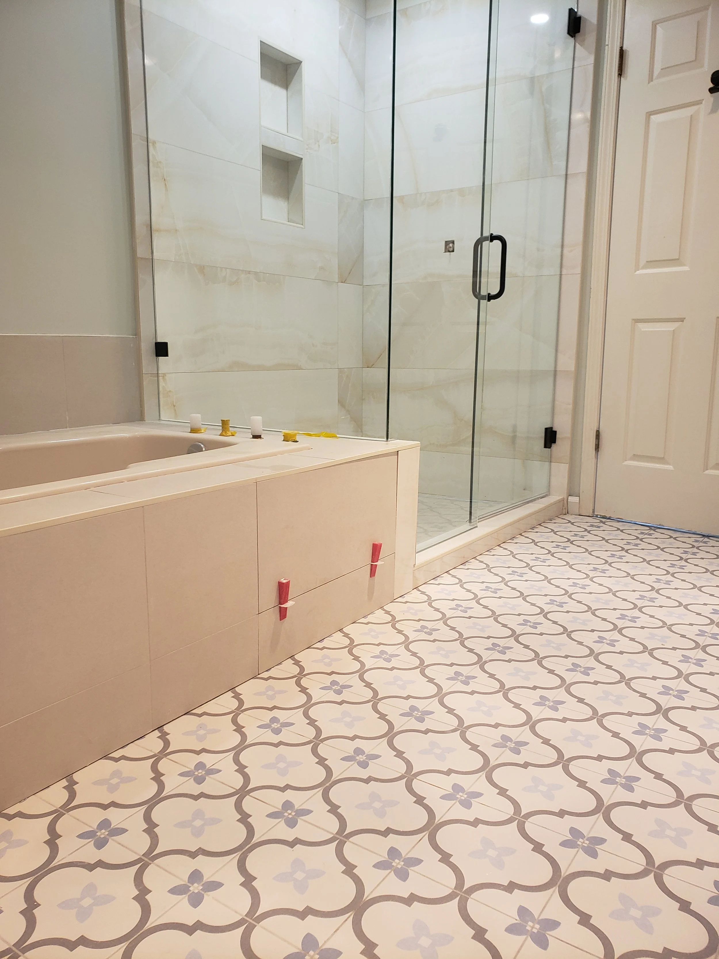 Bathroom with a bathtub on the left and a glass shower enclosure on the right, marble walls, patterned tile floor, and a closed white door.