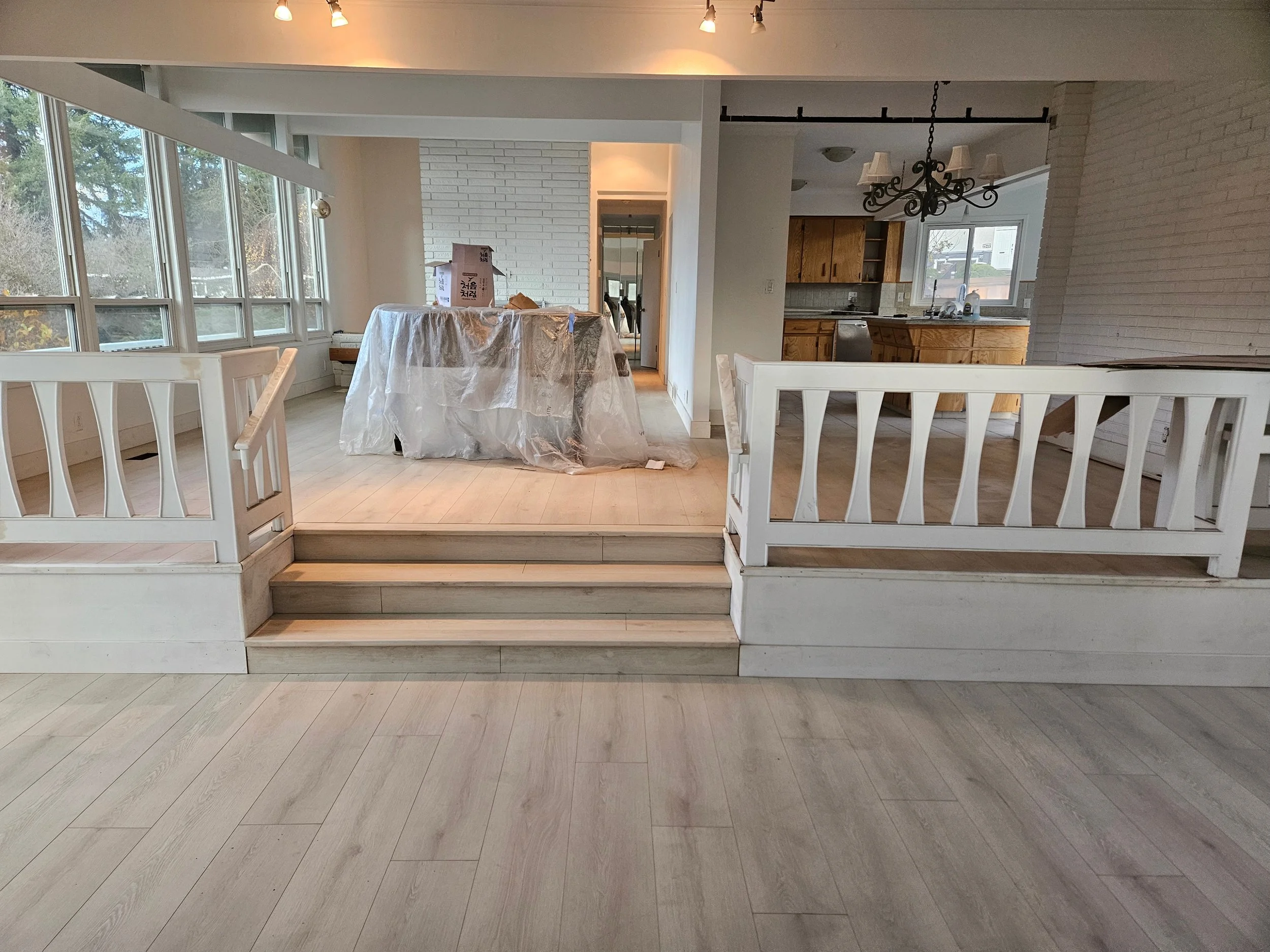Empty house interior with light wood flooring, white staircase with railing, and a kitchen with wooden cabinets. A covered table is visible, indicating ongoing renovation or furniture moving.