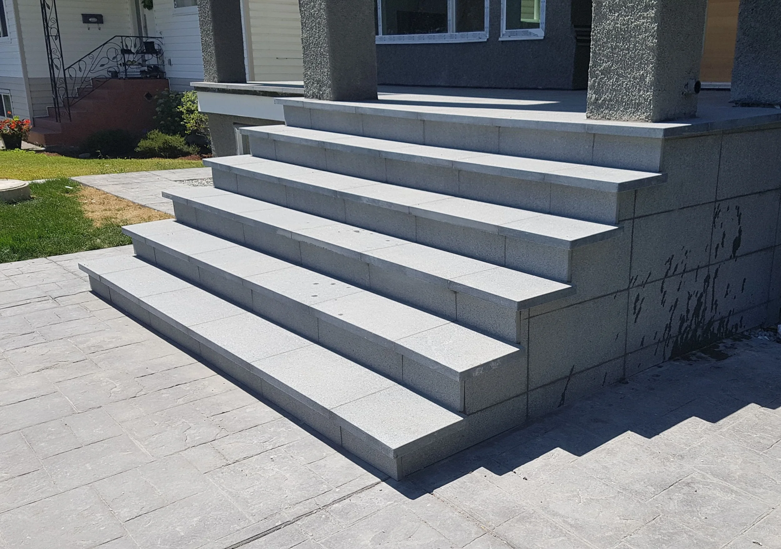 Concrete stairs leading up to a house entrance, with a green lawn and part of another house visible in the background.