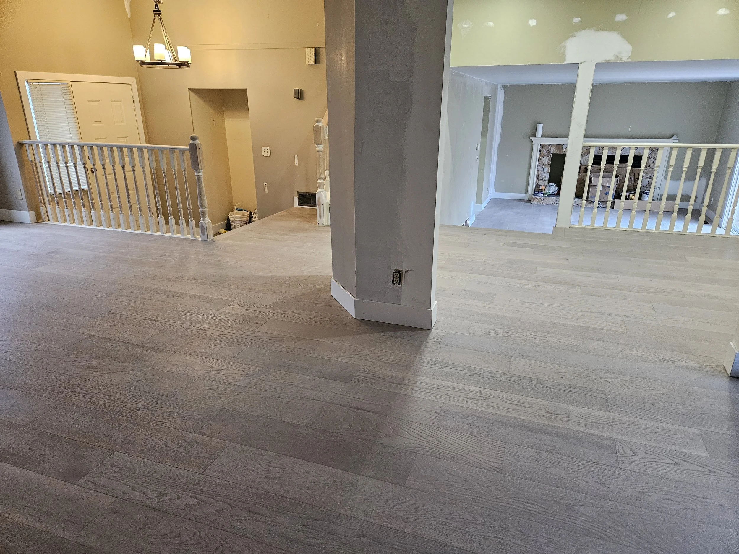 Interior view of a house with hardwood floors, white railings, and a staircase leading to the lower level. The walls are painted in neutral colors, and there is a chandelier hanging from the ceiling. Some areas are under construction or renovation.
