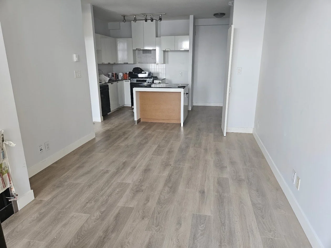 Empty living room with light-colored wood flooring, white walls, and a view into a kitchen with white cabinets and black appliances.