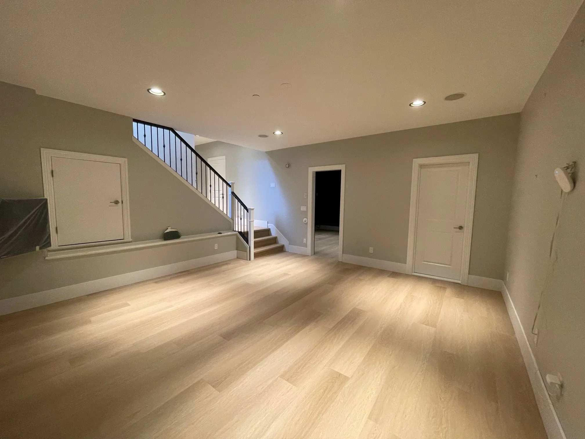 Empty room with light wood flooring, white trim, gray walls, recessed lighting, with a staircase leading upstairs on the left, and two closed white doors on the right. There is a small hatch door under the stairs and some transparent plastic covering part of a wall, possibly indicating ongoing work.
