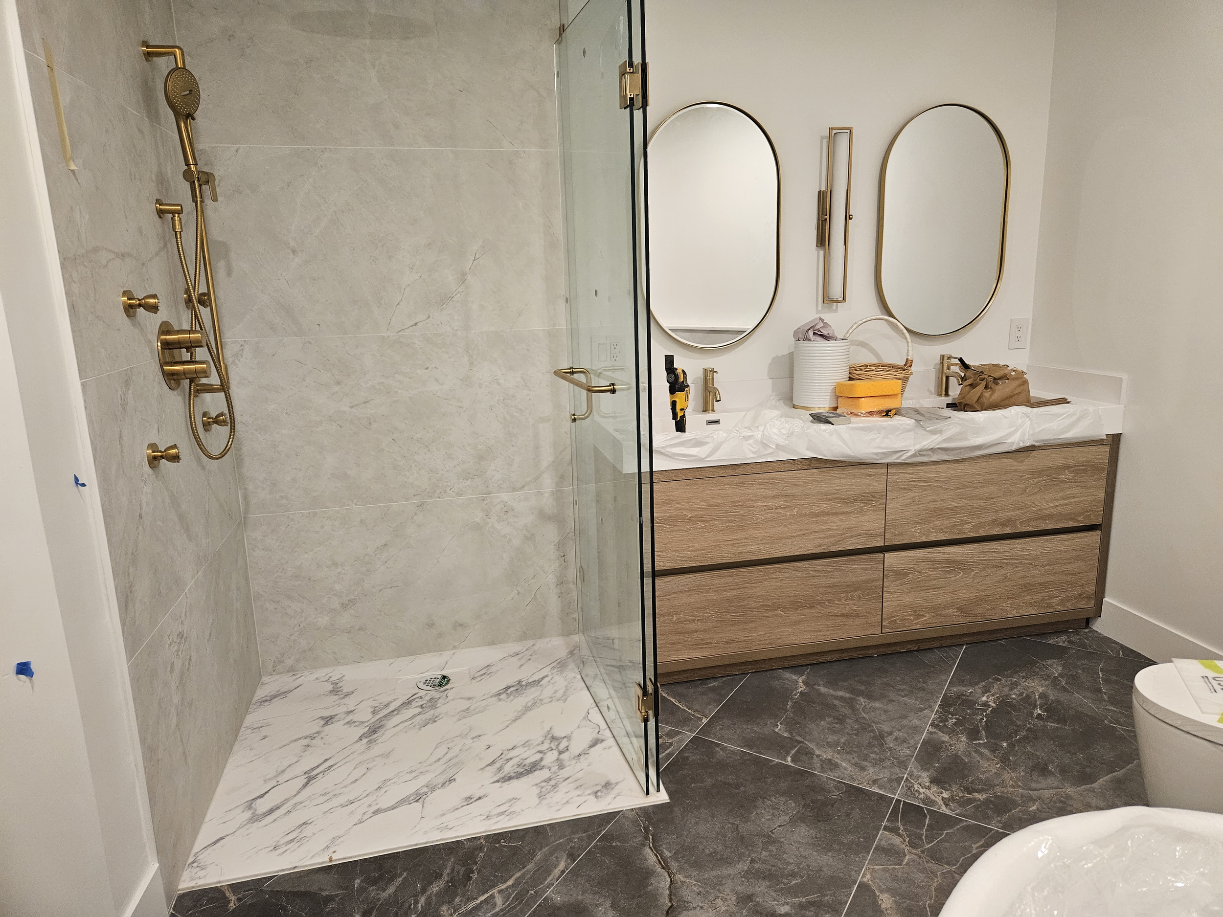 Modern bathroom with a walk-in shower featuring gold fixtures and marble tiles, a wooden vanity with two oval mirrors, and a marble floor.