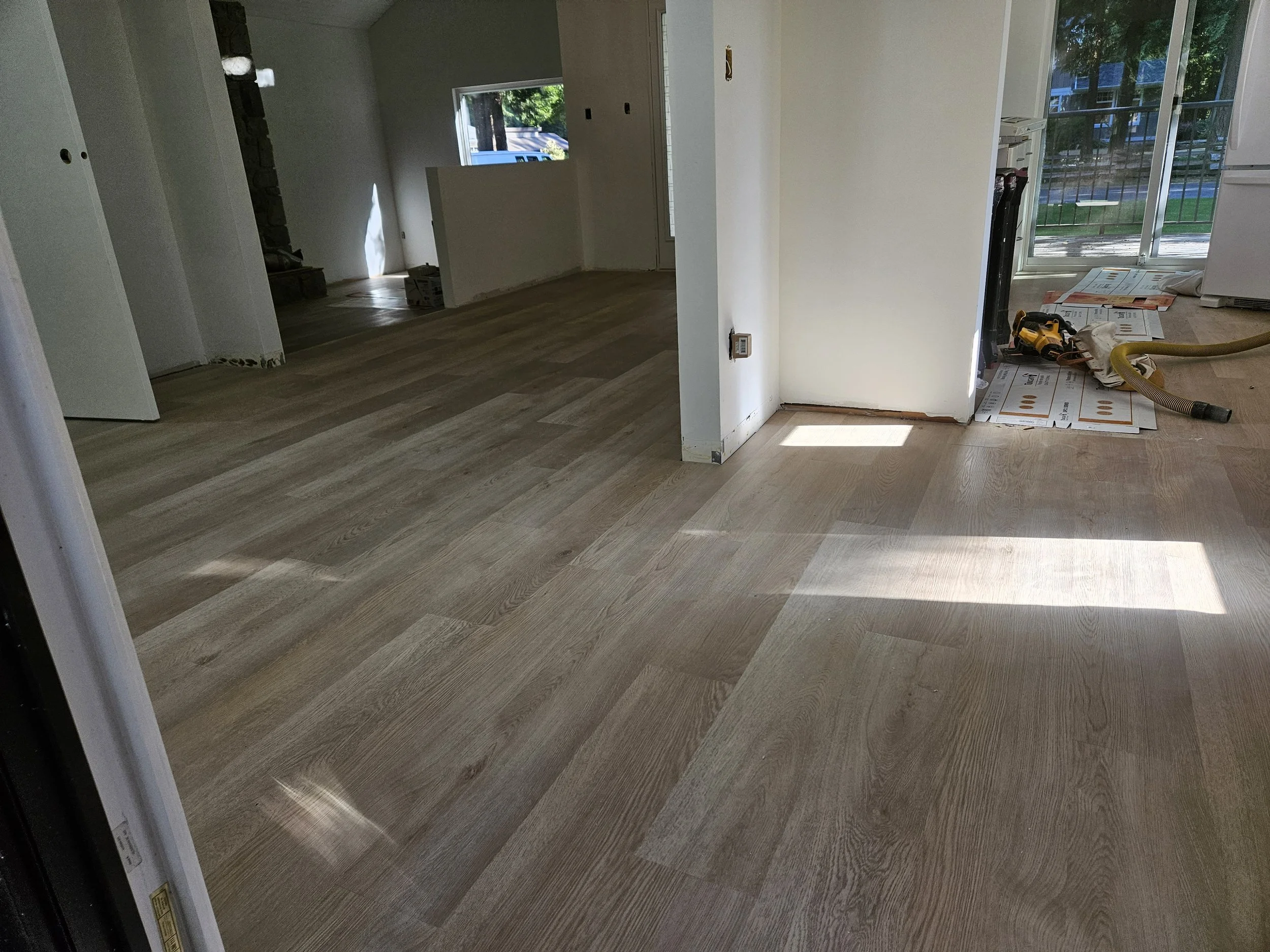 Interior of a house under renovation with new hardwood flooring, partially installed, near sliding glass doors leading outside with a patio and lawn.