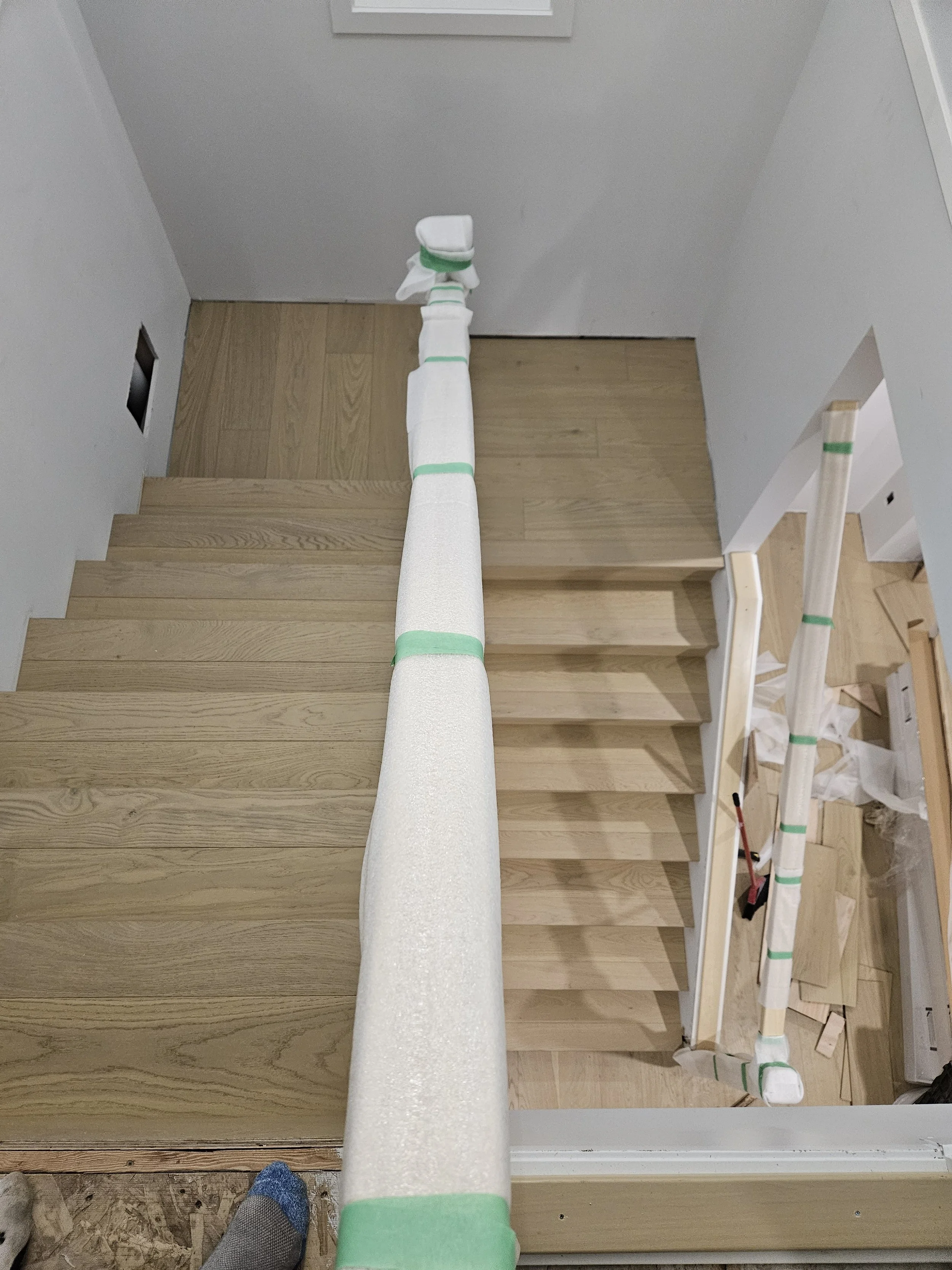 View of a staircase under construction with a temporary protective covering on the handrail, partially covered stairs, an unfinished wall, and tools on the floor.