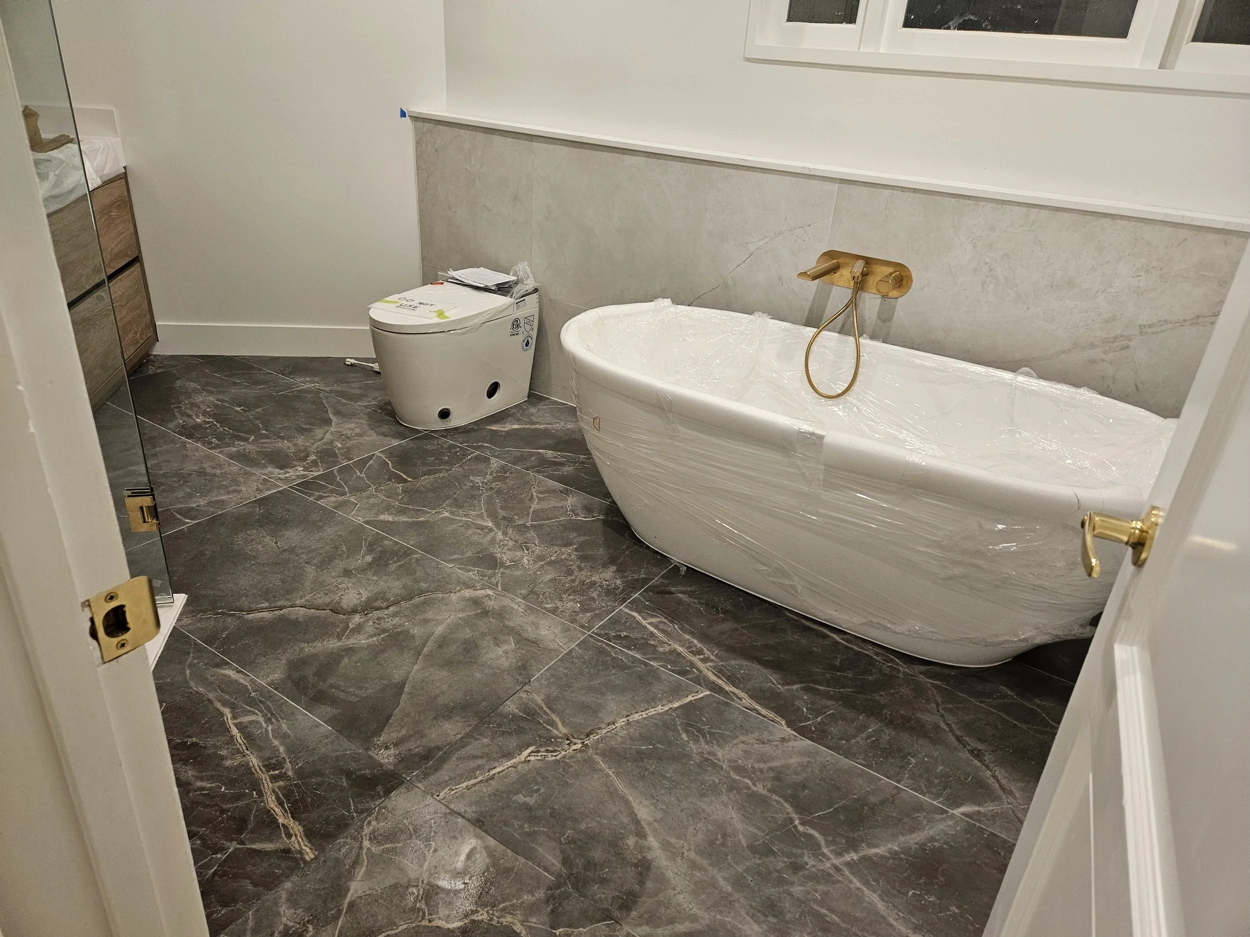 A bathroom with a marble floor, a white bathtub covered in plastic, a toilet with a paper on top, and a vanity with drawers. The wall behind the bathtub is light-colored, and there is a window above the bathtub.