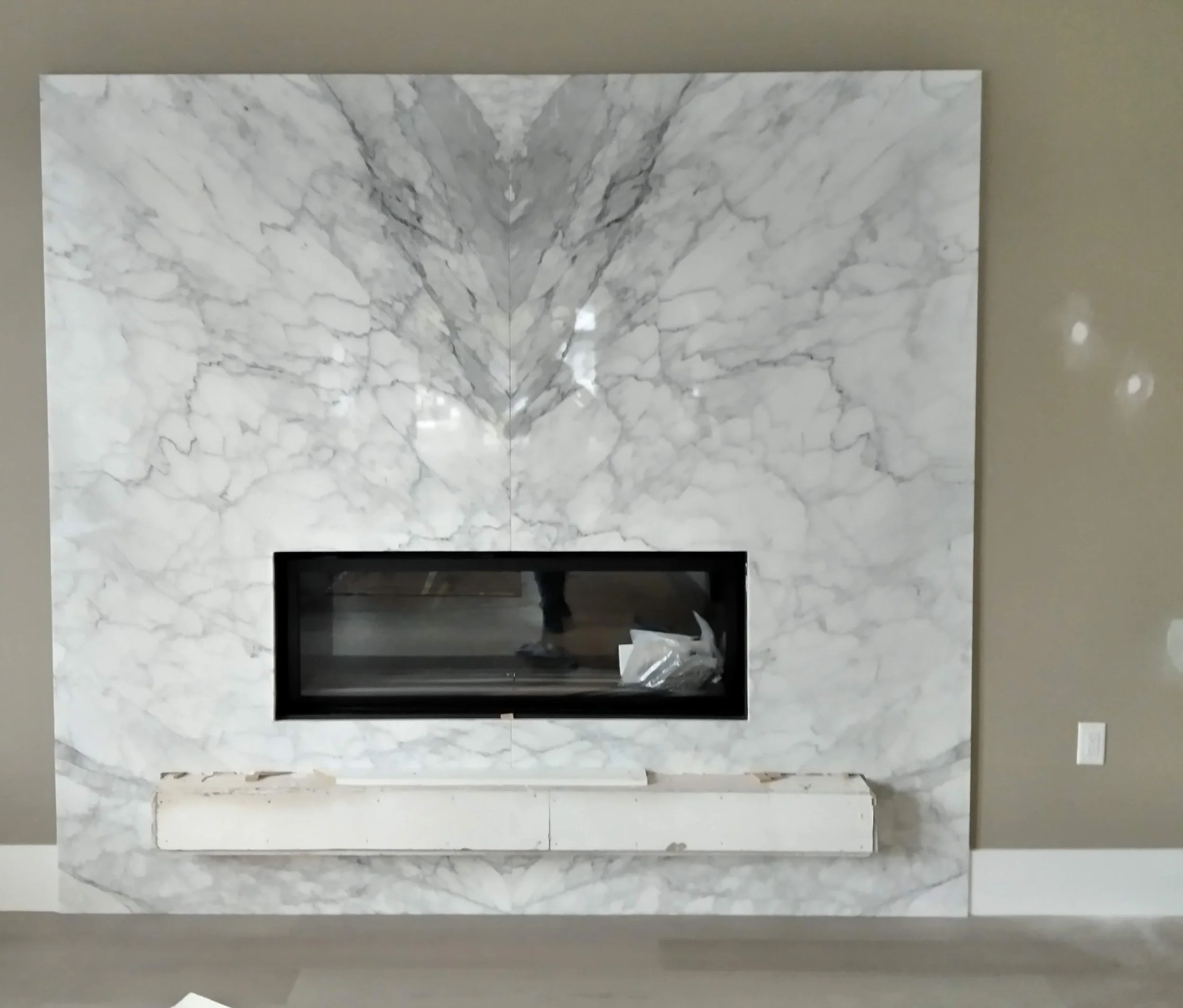 A modern fireplace with a black glass insert set in a large white marble wall with gray veining, and a white marble mantel in front.