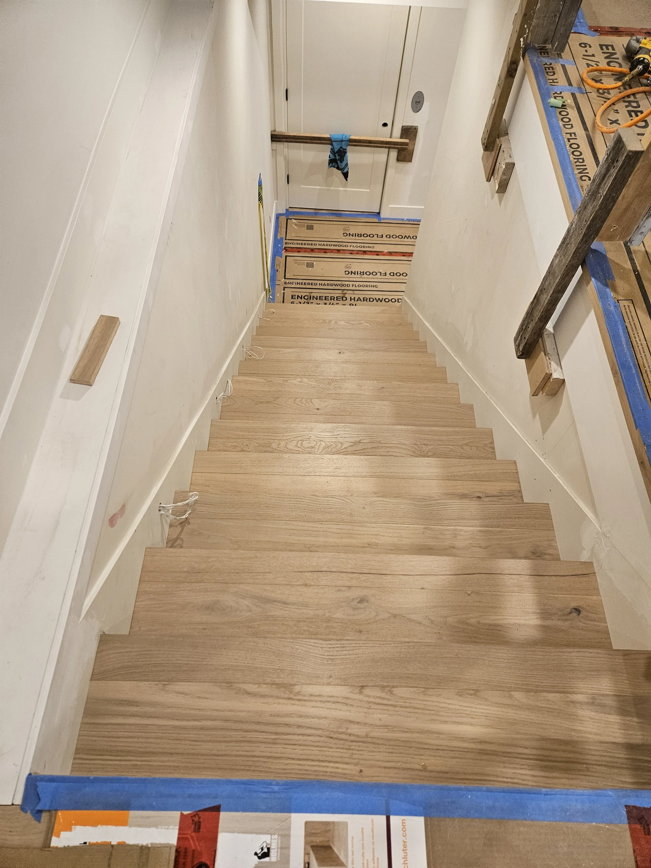 Wooden staircase under construction with protective blue tape at the bottom, and construction materials and tools visible around, including a workbench with orange scissors and extension cord.