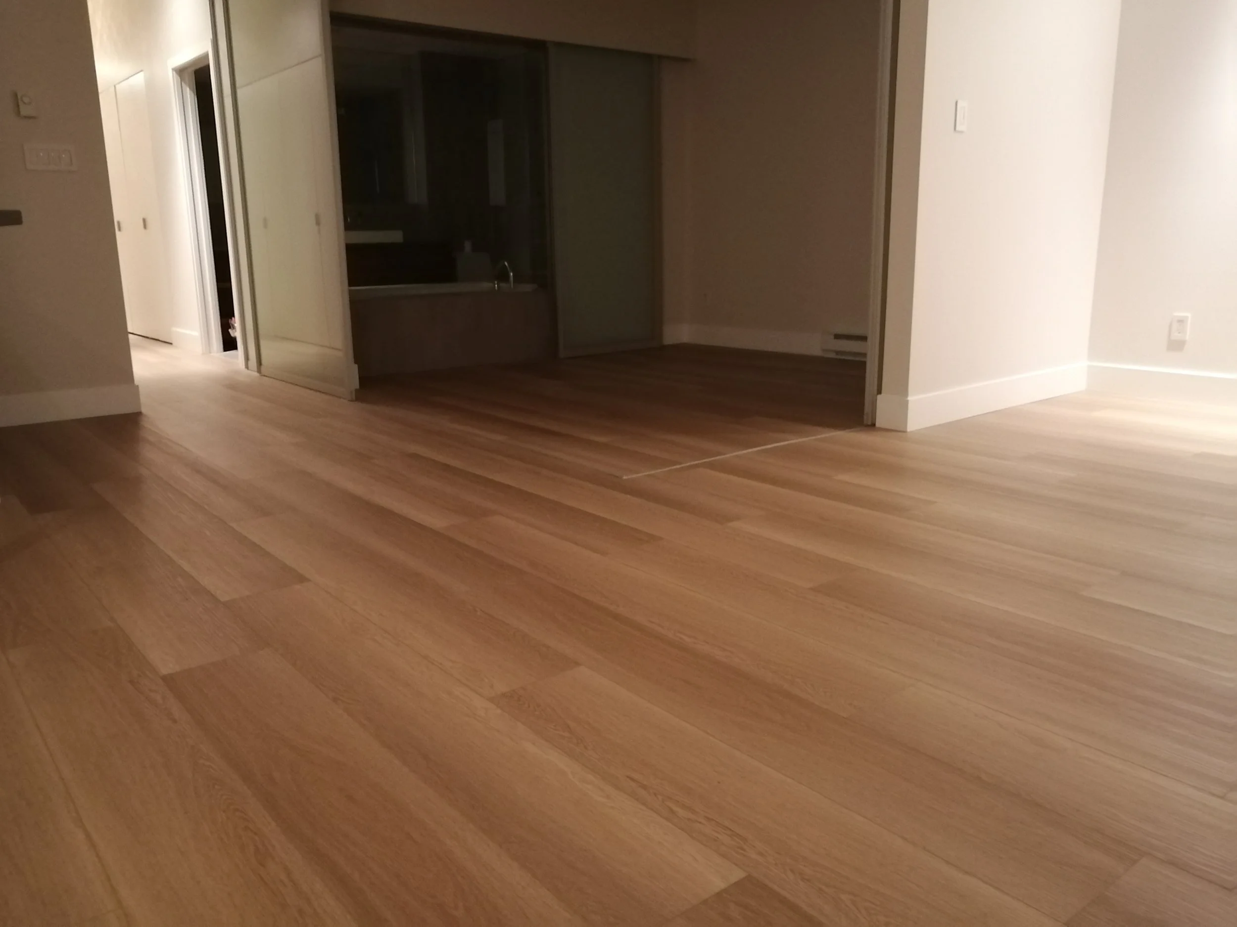 Empty living space with wooden flooring, partially open sliding glass doors leading to a kitchen, and white walls.