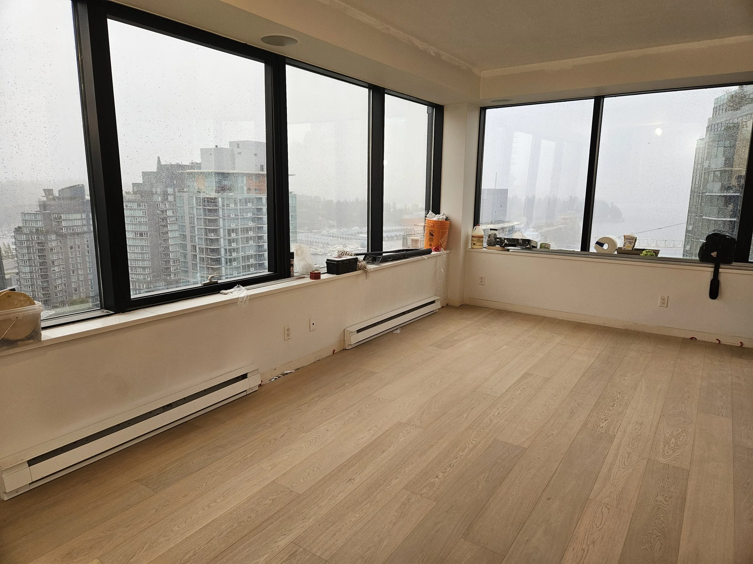 Empty room with large windows overlooking a cityscape of high-rise buildings on a rainy day.