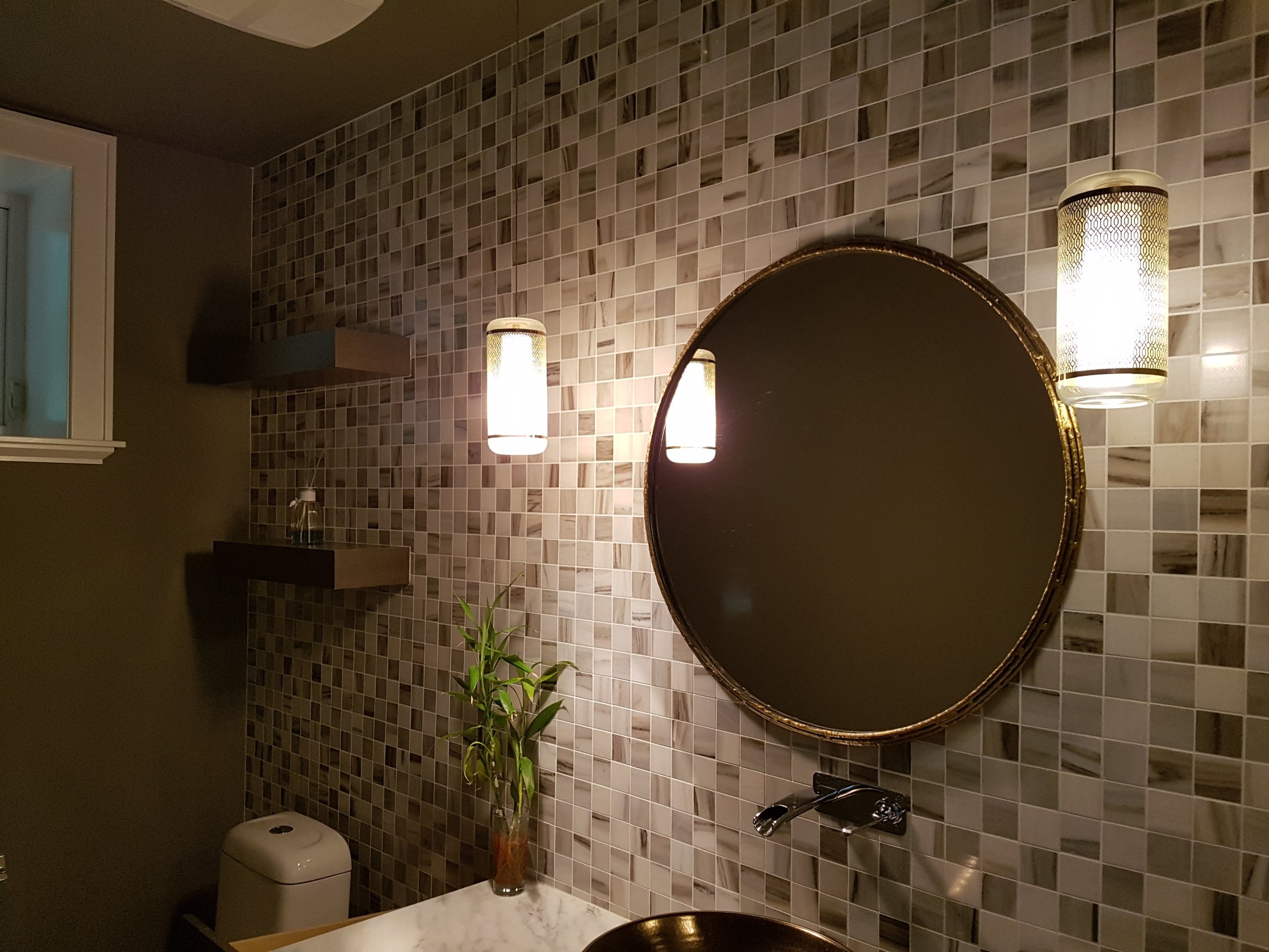 Bathroom with round mirror, two hanging lights, tiled wall, small window, marble countertop, green plant, and a toilet.