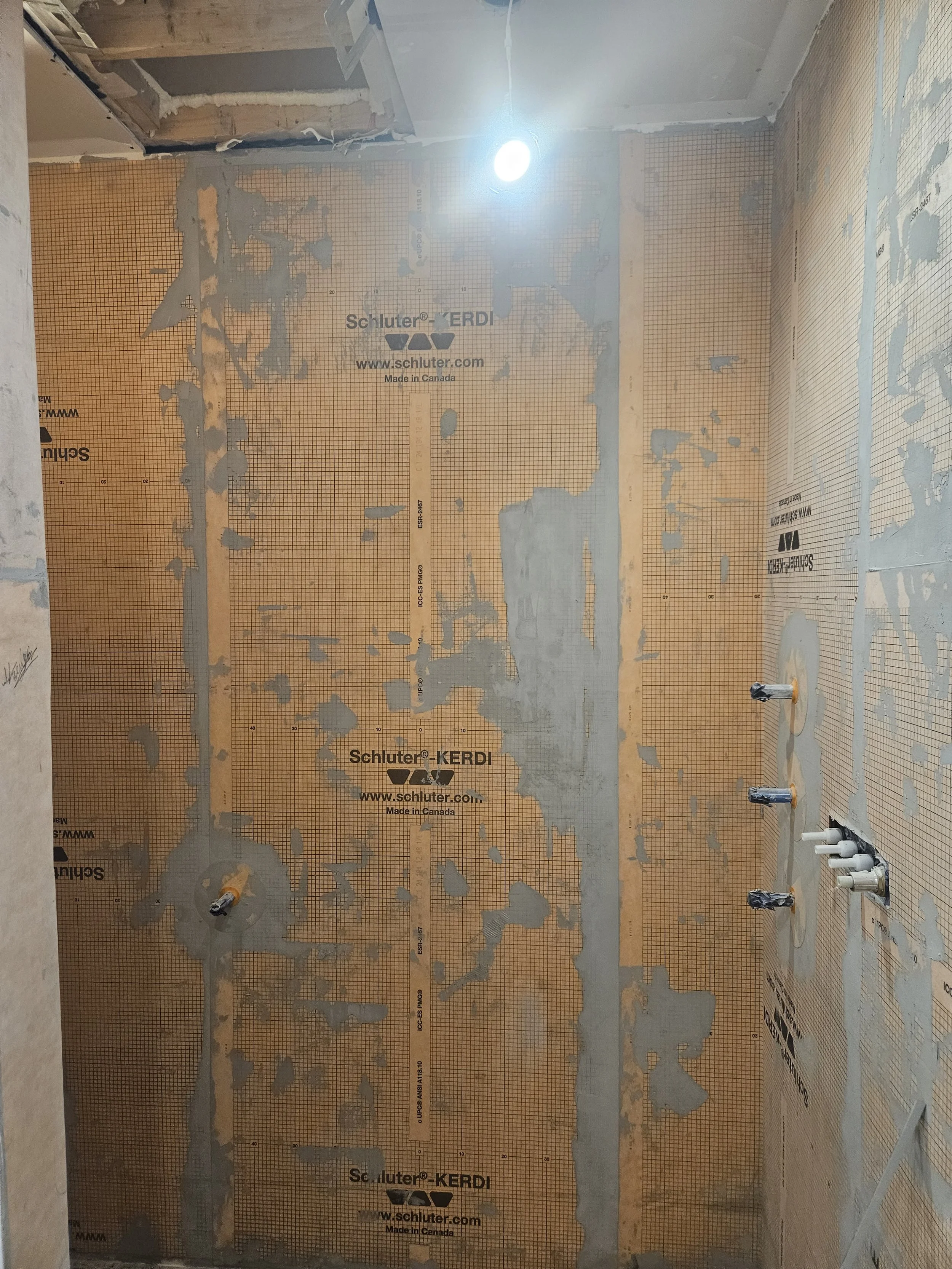 Bathroom wall under construction with plumbing pipes, drywall, and insulation material, with a light bulb overhead.