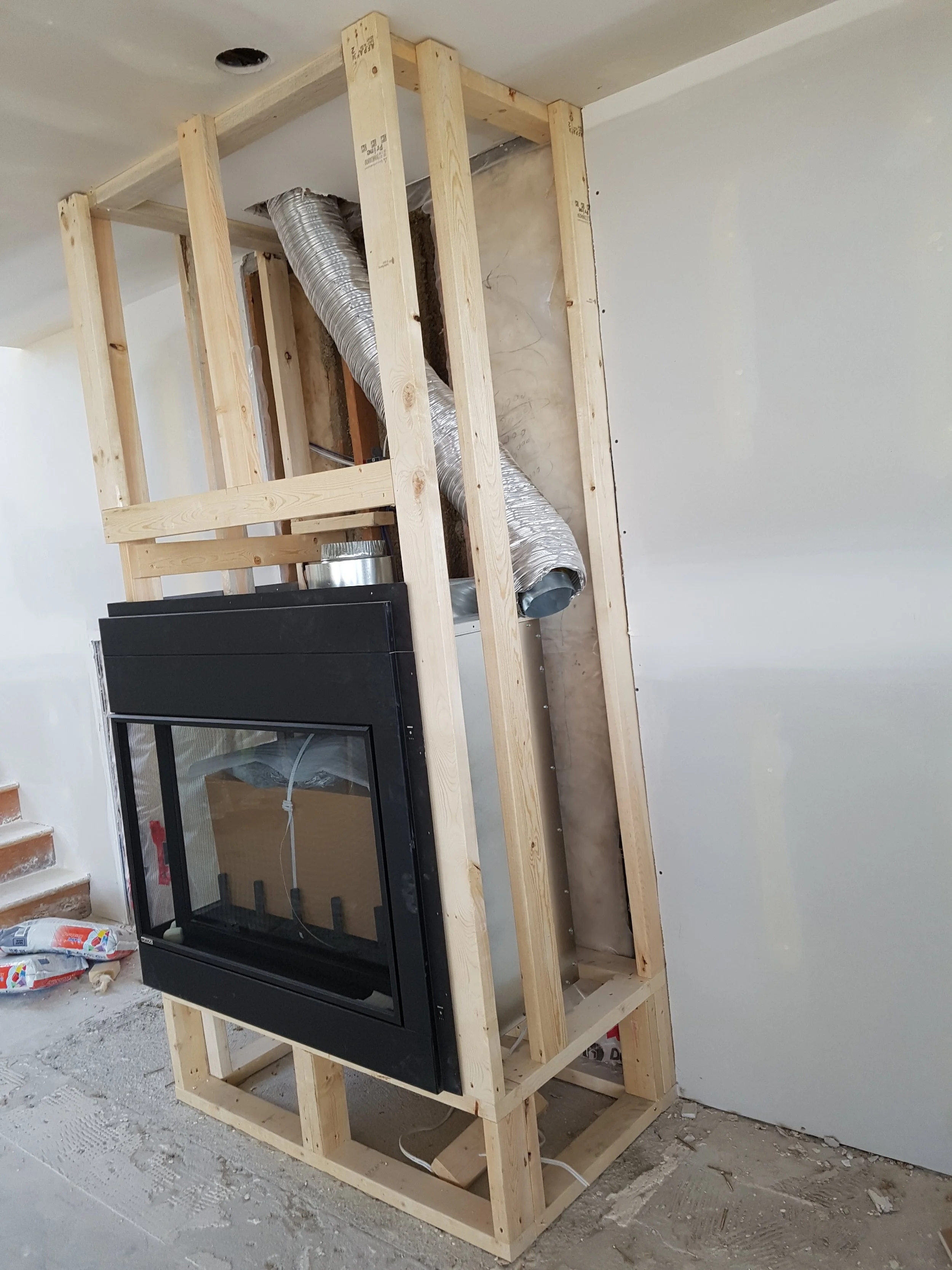 Construction of a fireplace surround in progress with wooden framing, a glass-front fireplace insert, and HVAC ductwork in a room under renovation.