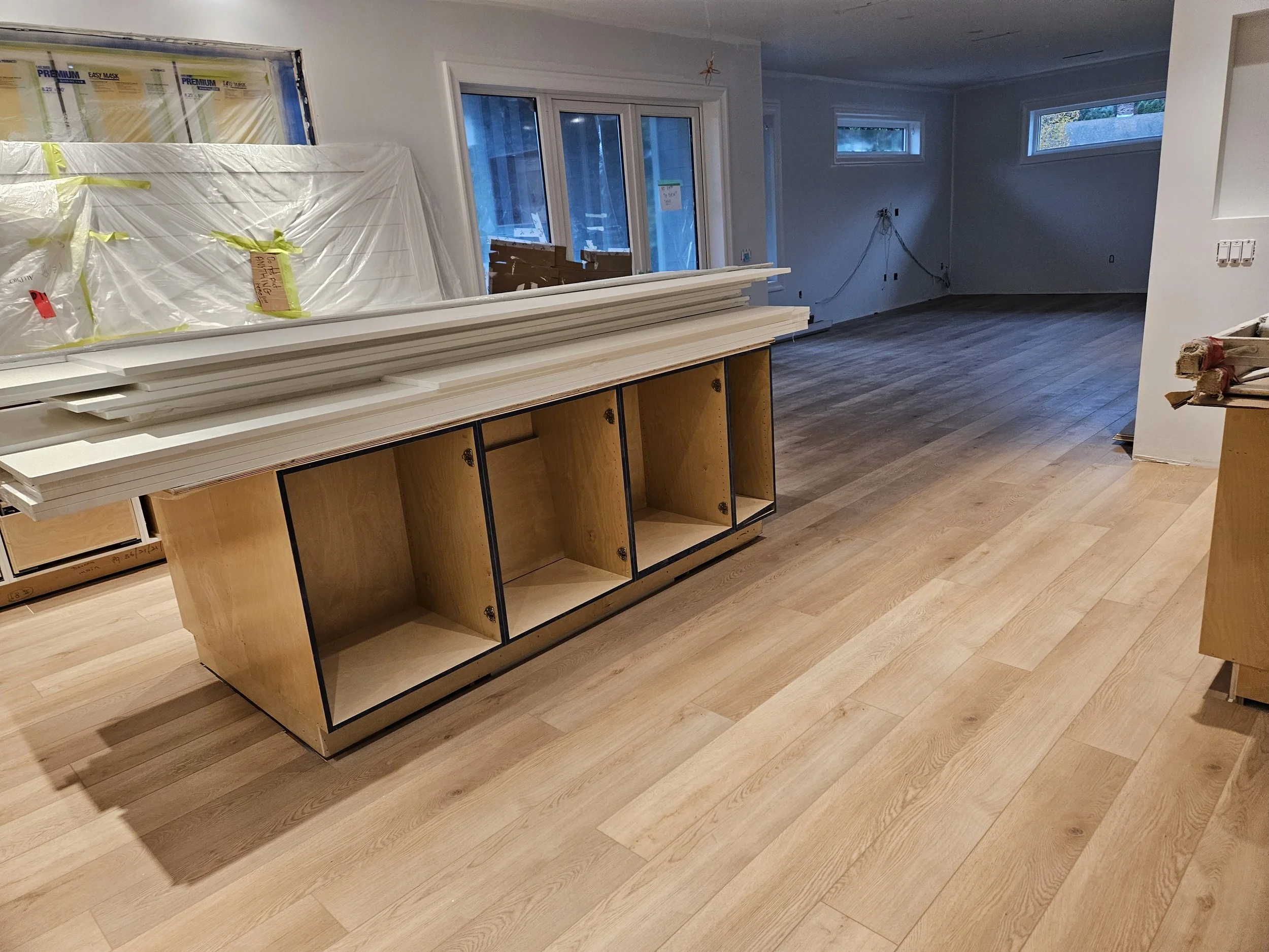 Interior of a house under renovation with wooden floors, a partially assembled cabinet, and construction materials. There are large windows and sliding glass doors, with some wrapped packages and tools visible.