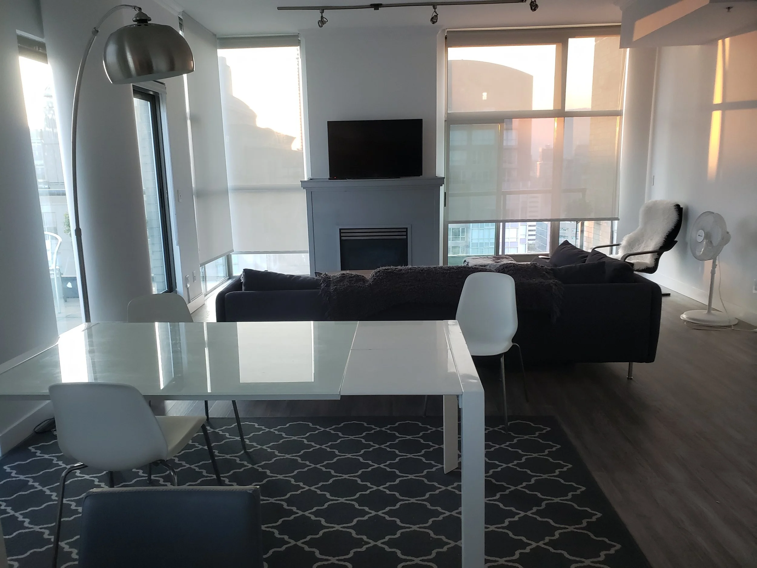 Modern living room with white dining table, white chairs, black sofa, tall silver floor lamp, fireplace, flat-screen TV, armchair, and standing fan.