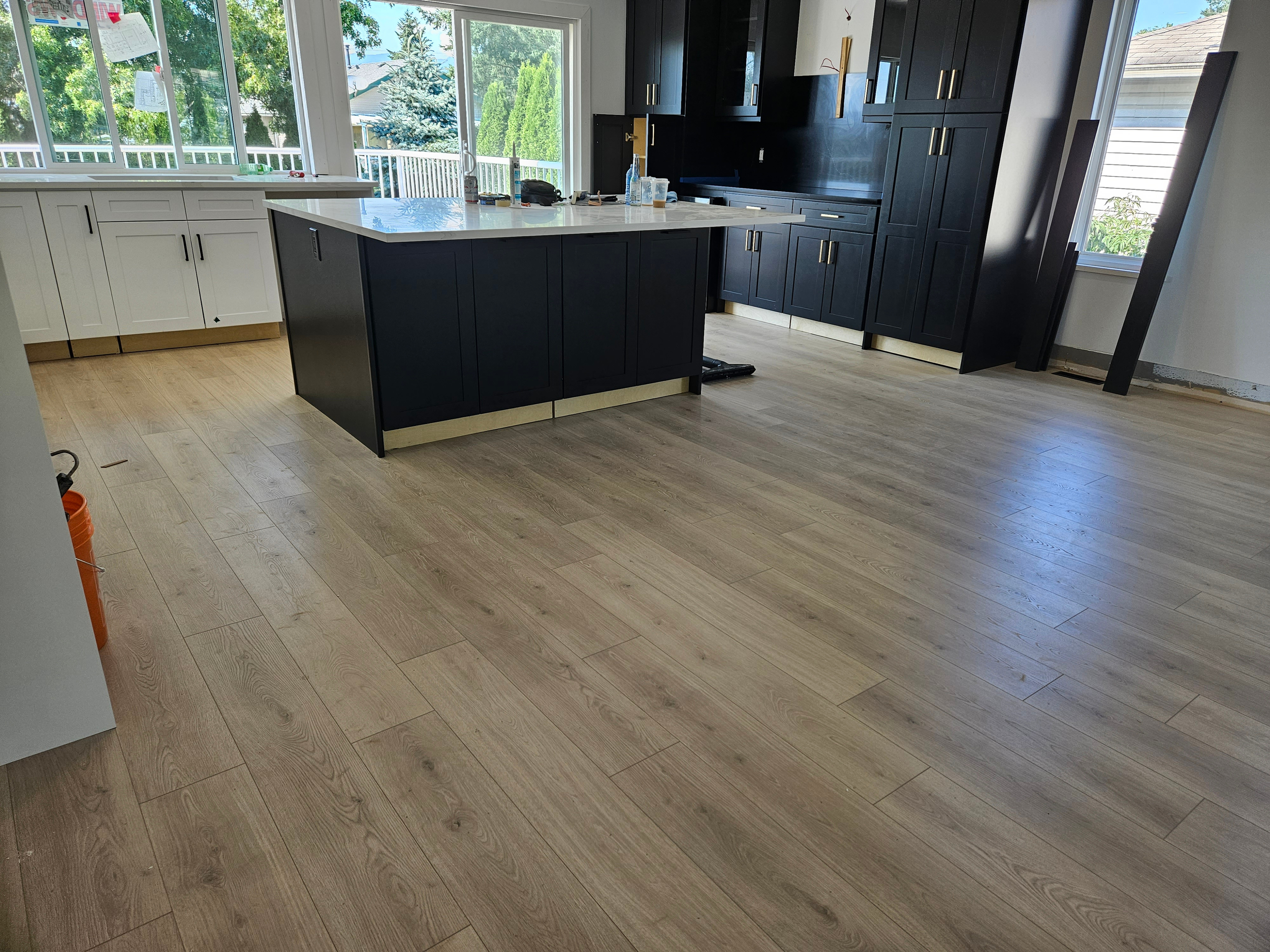 Open kitchen with light-colored wood flooring, white cabinets, black kitchen island, and large windows showing trees outside.