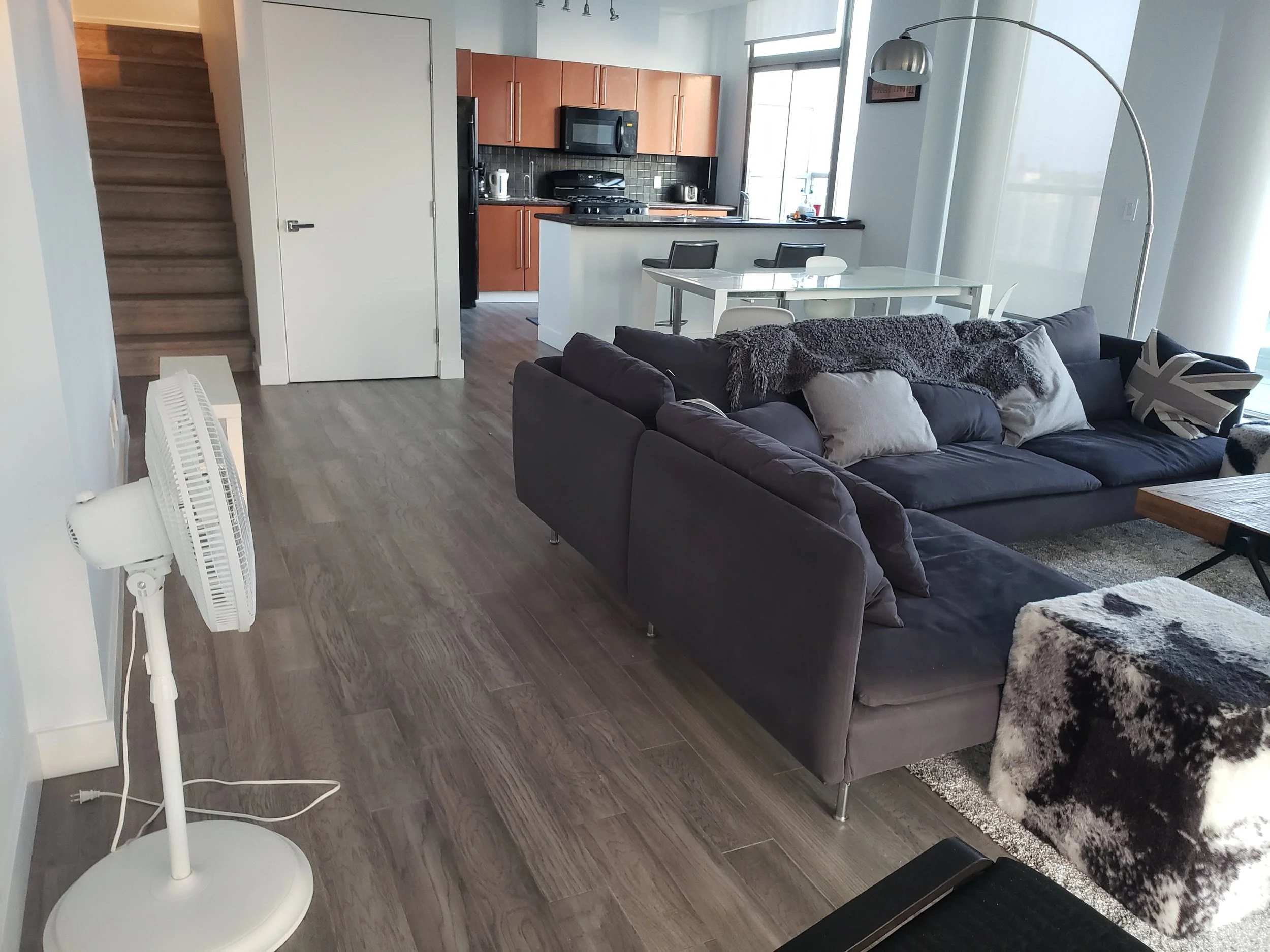 Living room with a dark couch, white pillows, a floor lamp, and a TV in the corner. Adjacent kitchen with wooden cabinets, black appliances, and a dining table with chairs. Light-colored hardwood floors, a fan, and large windows letting in natural light.
