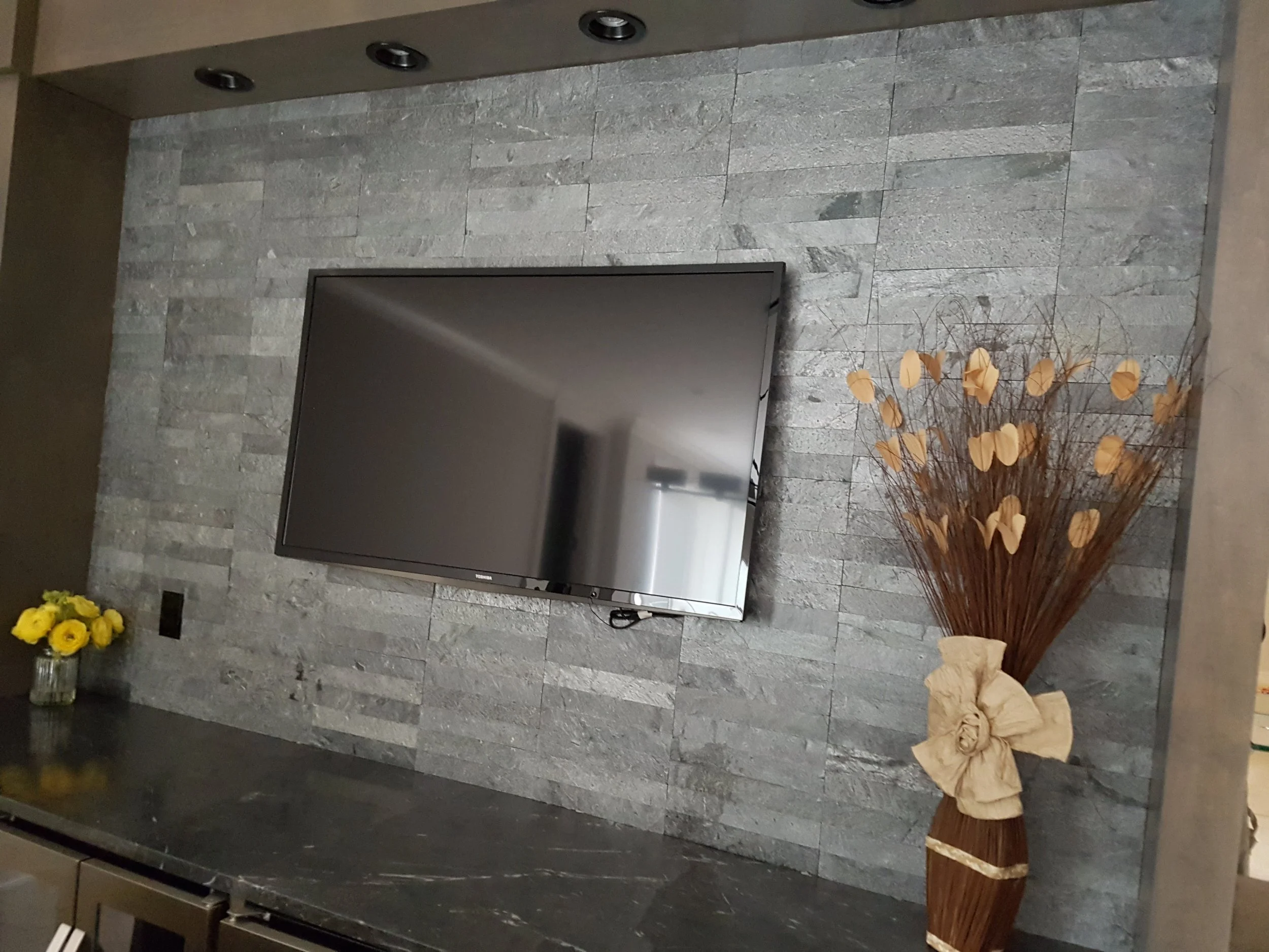 A wall with gray stone tiles, a flat-screen television mounted on the wall, a small bouquet of yellow flowers in a glass jar on a dark surface, and a large decorative vase with dried brown twigs and beige circular ornaments.