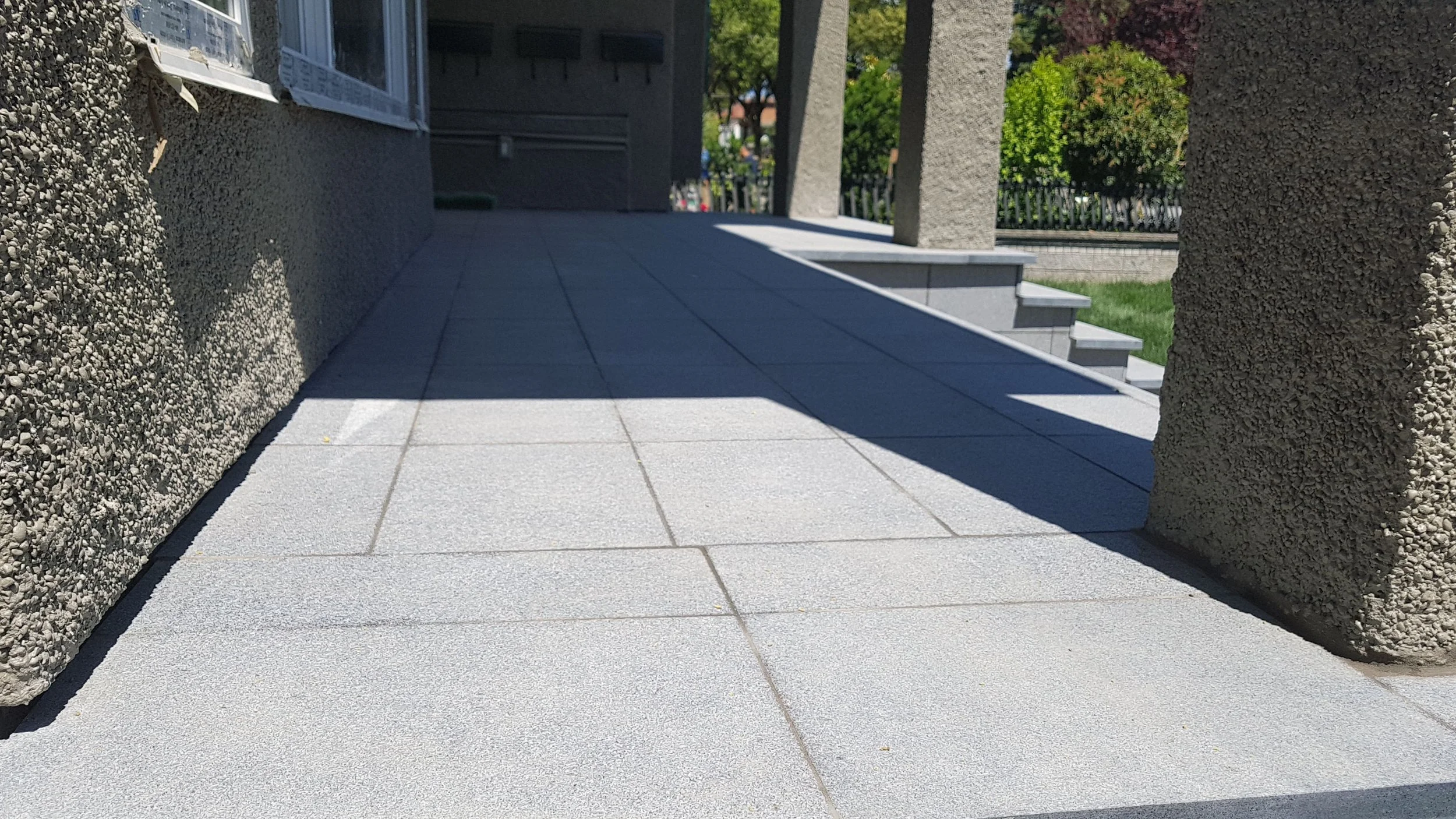 A close-up of a concrete tiled walkway next to a building with textured exterior walls, with stairs and a green yard in the background.