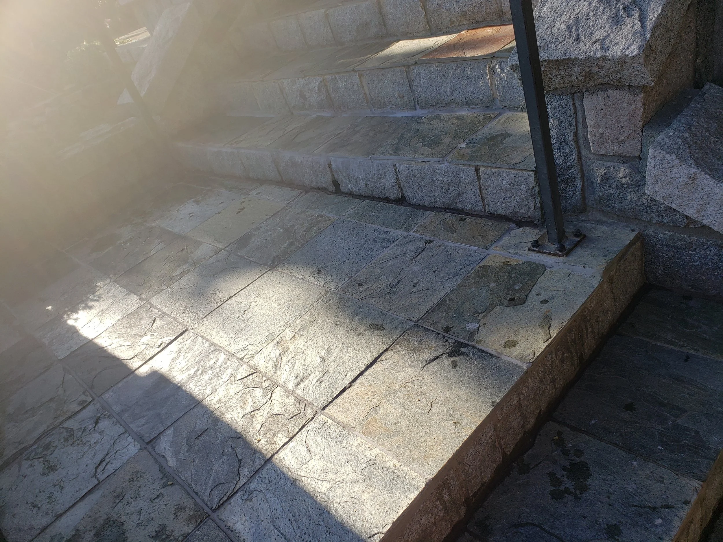Close-up of a stone-tiled outdoor patio with stairs and a black metal railing, sunlight casting shadows on the surface.