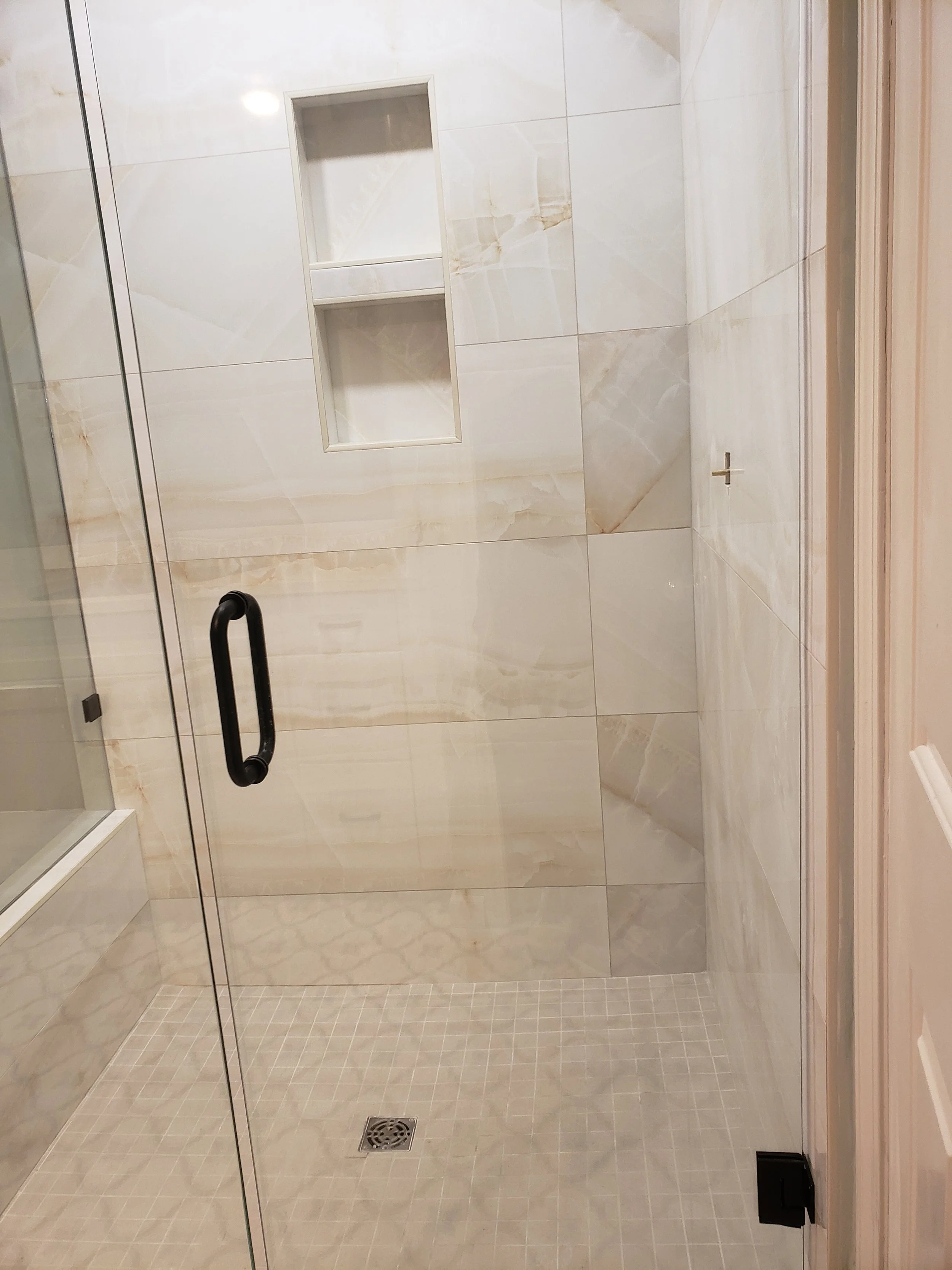 Empty walk-in shower with beige marble tiles, a built-in niche with two shelves, a black handle on the glass door, and a floor drain.