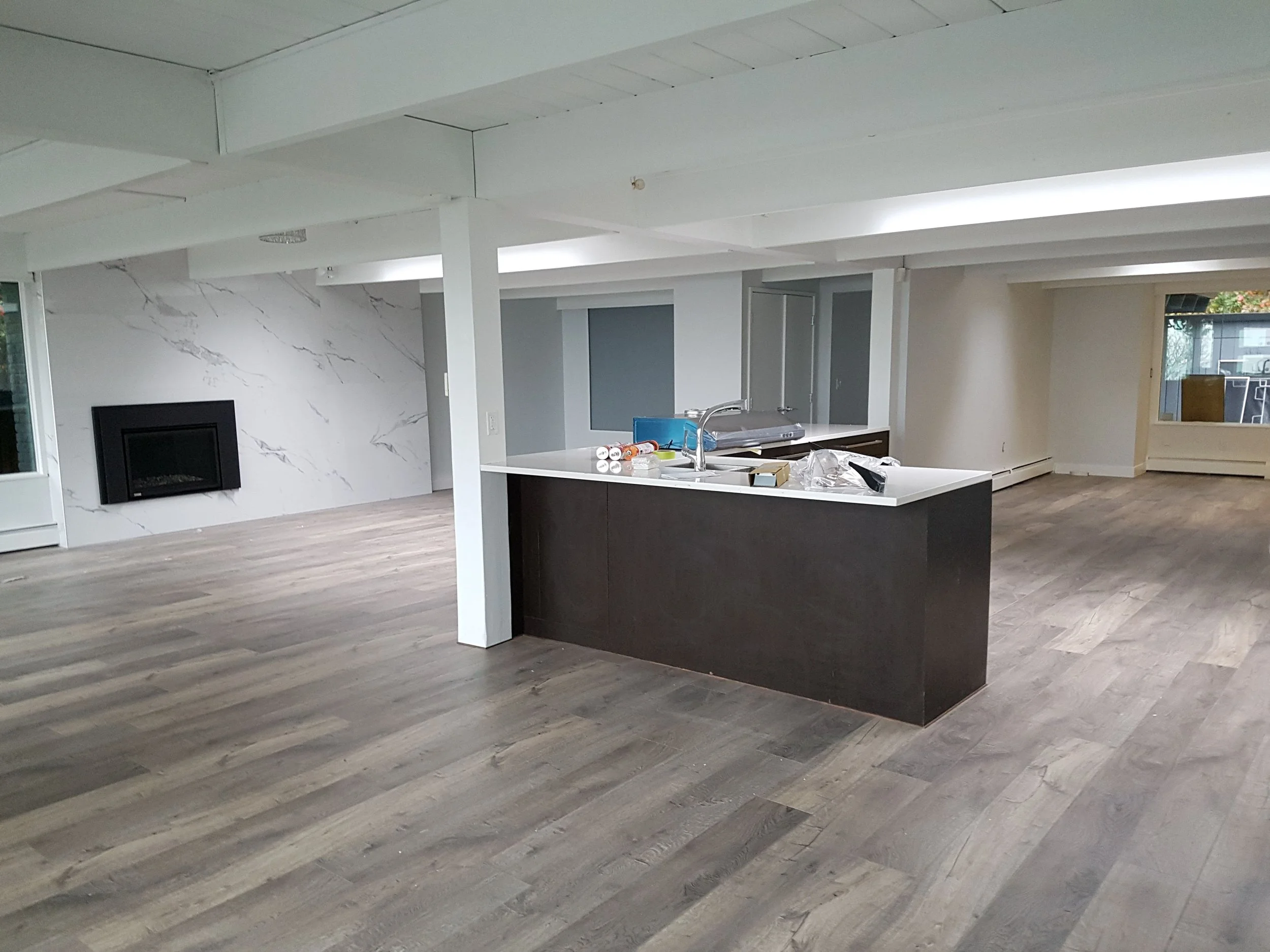 Empty modern living space with hardwood floors, a white marble accent wall, and a built-in fireplace, with a kitchen island in the foreground.