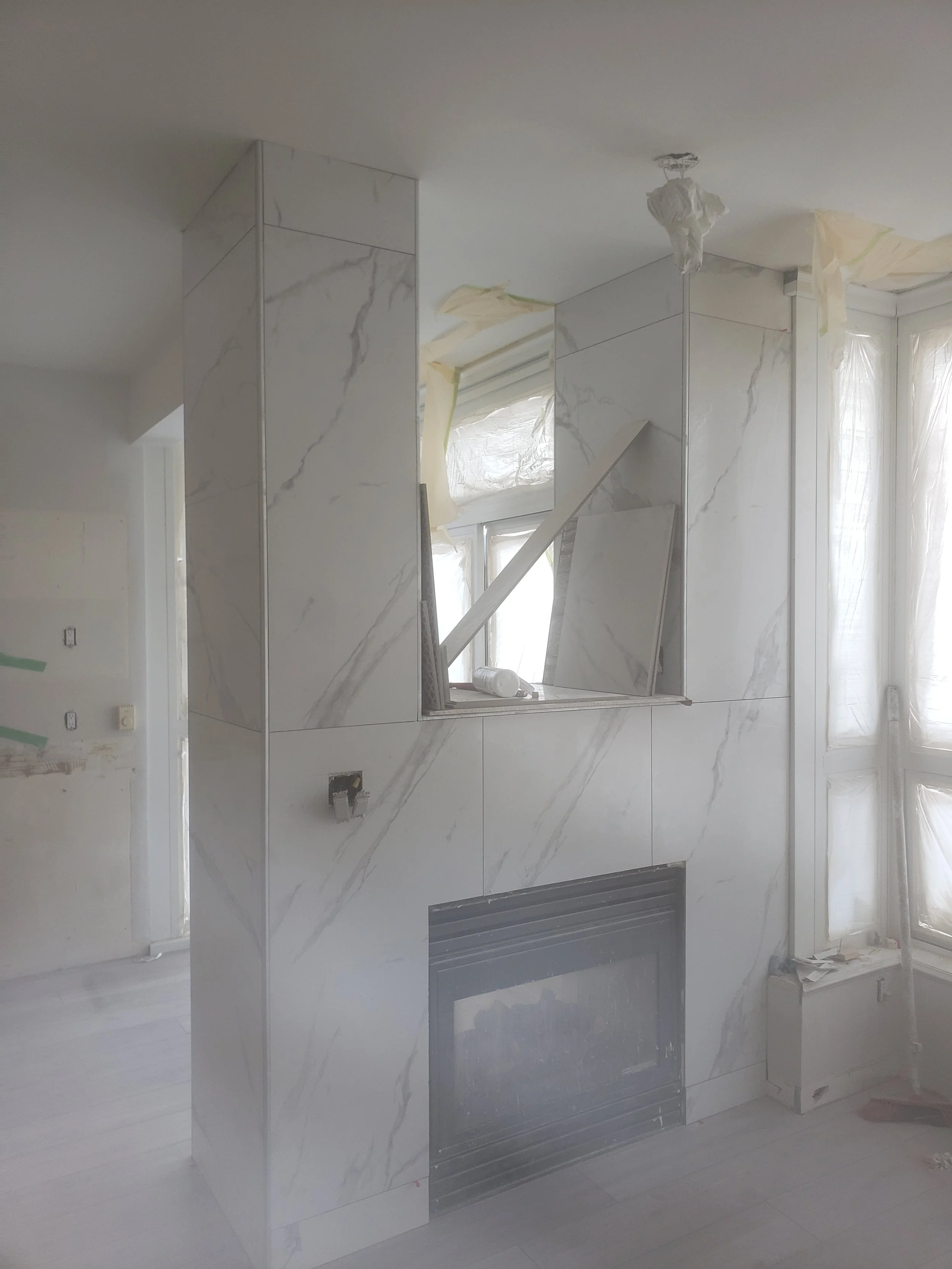 A partially finished room featuring a marble fireplace surround and chimney with a rectangular opening, newer window with plastic coverings, and construction materials around, in a space under renovation.