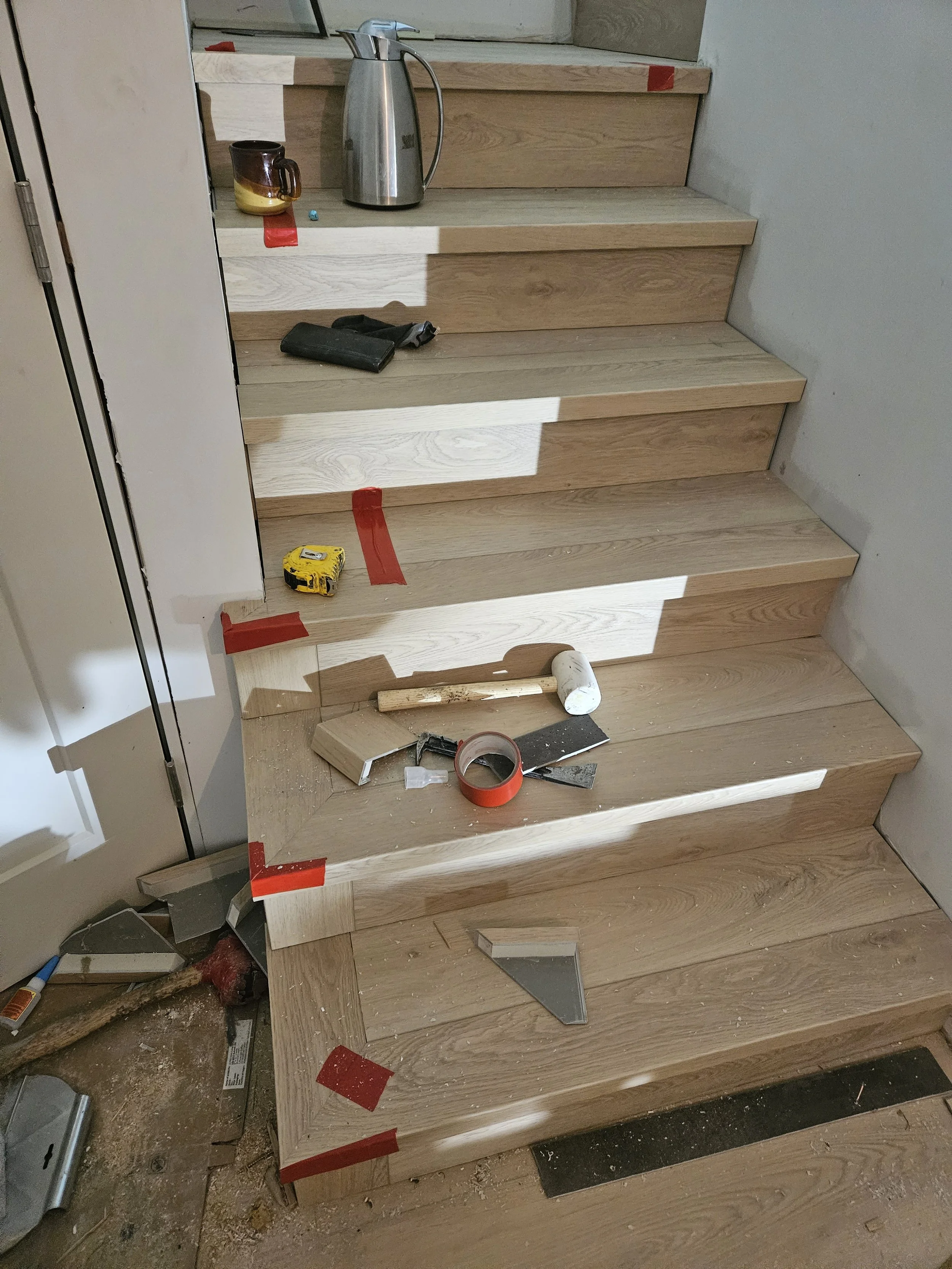 Construction tools and materials on wooden stairs, including a hammer, tape measure, and tiles, with red tape marking sections, and a water kettle and cup on the landing.