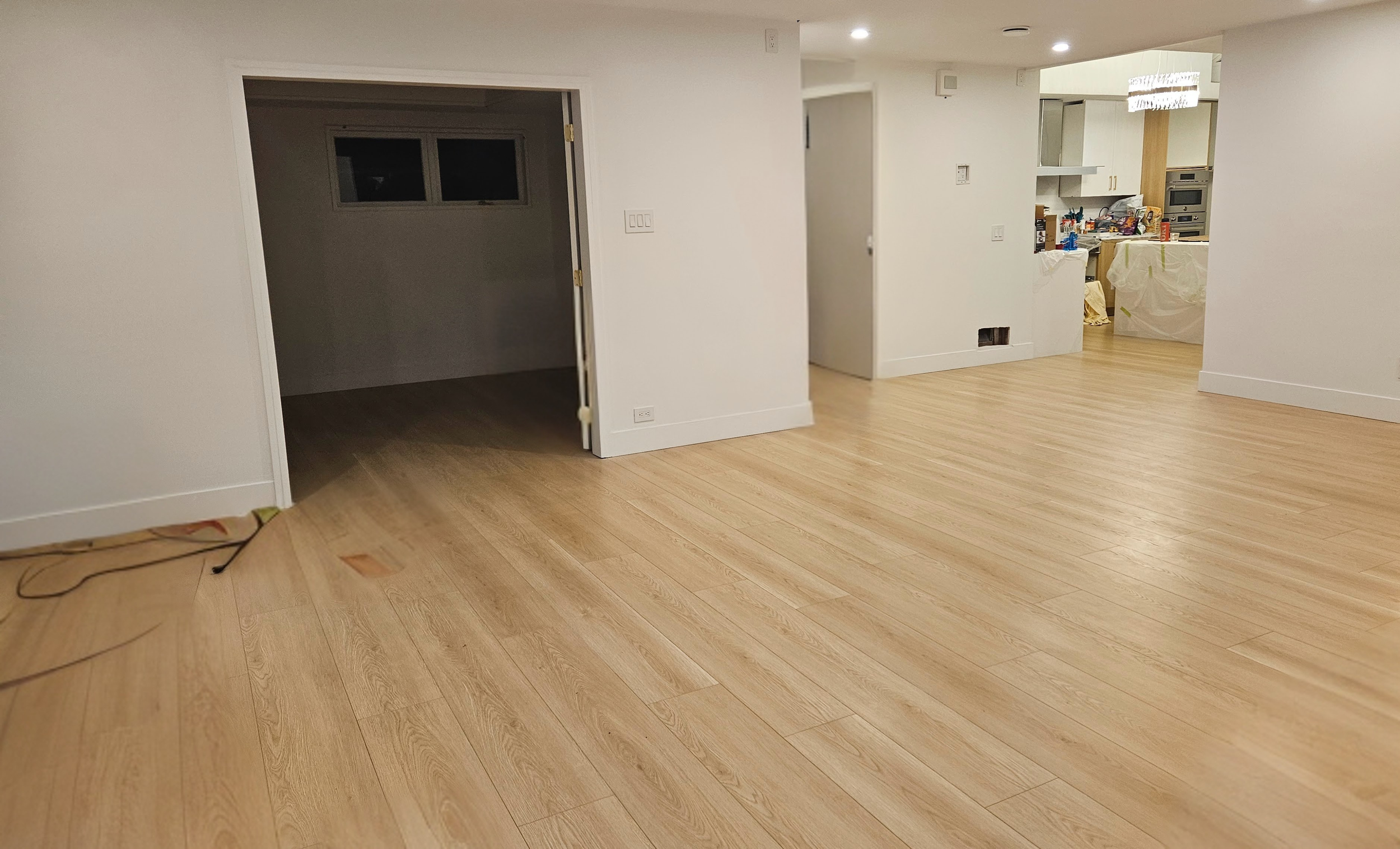 Empty living room with light wood flooring, white walls, and visible kitchen in the background; a dark oval opening leads to another room, and a small window is on the left wall.