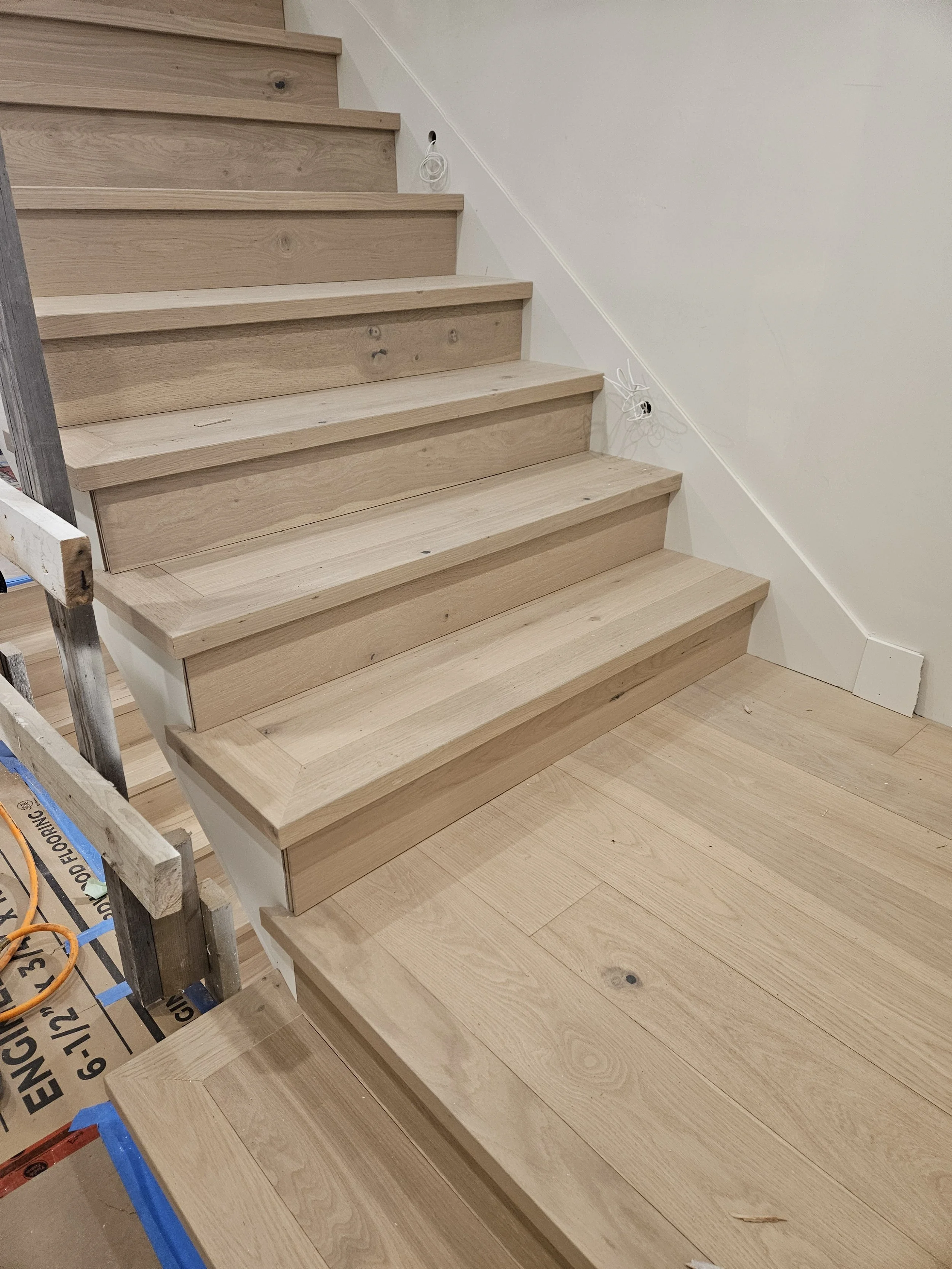 The image shows a stairway under construction with wooden steps and flooring, along with unfinished drywall walls and visible electrical wiring.