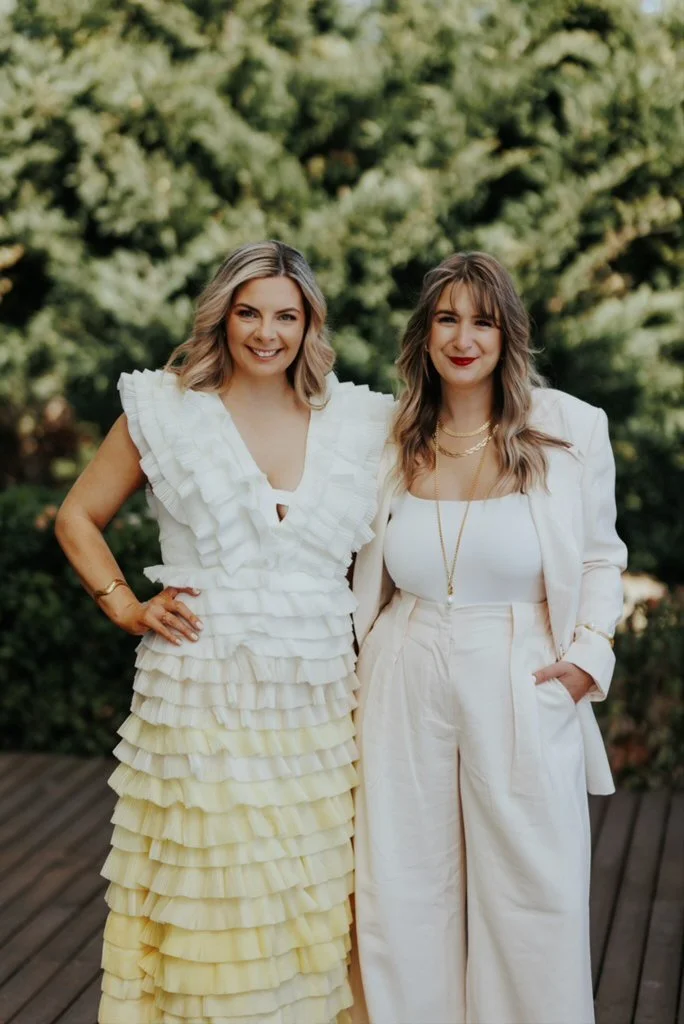 Two women standing together outdoors, smiling, with a blurred green leafy background. One woman wears a white ruffled dress with layered fabric, and the other wears a white suit with a tank top and gold jewelry.