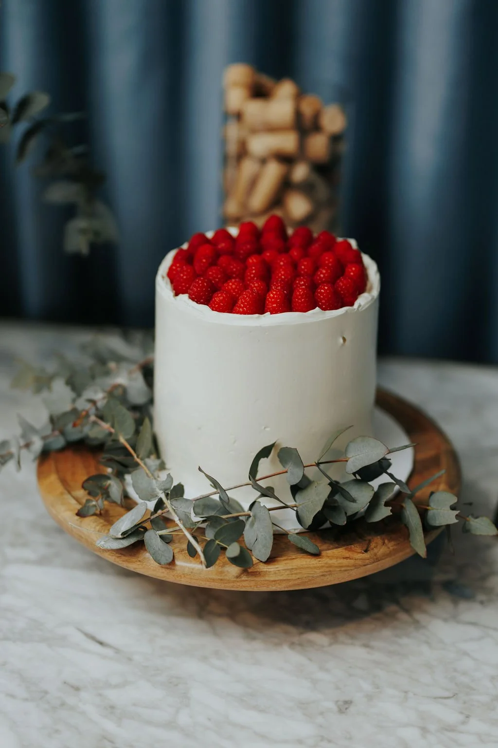 A white frosted cake topped with fresh red raspberries, placed on a round wooden serving board with eucalyptus leaves around the base. In the background, there is a bundle of chopped firewood and a blue curtain.