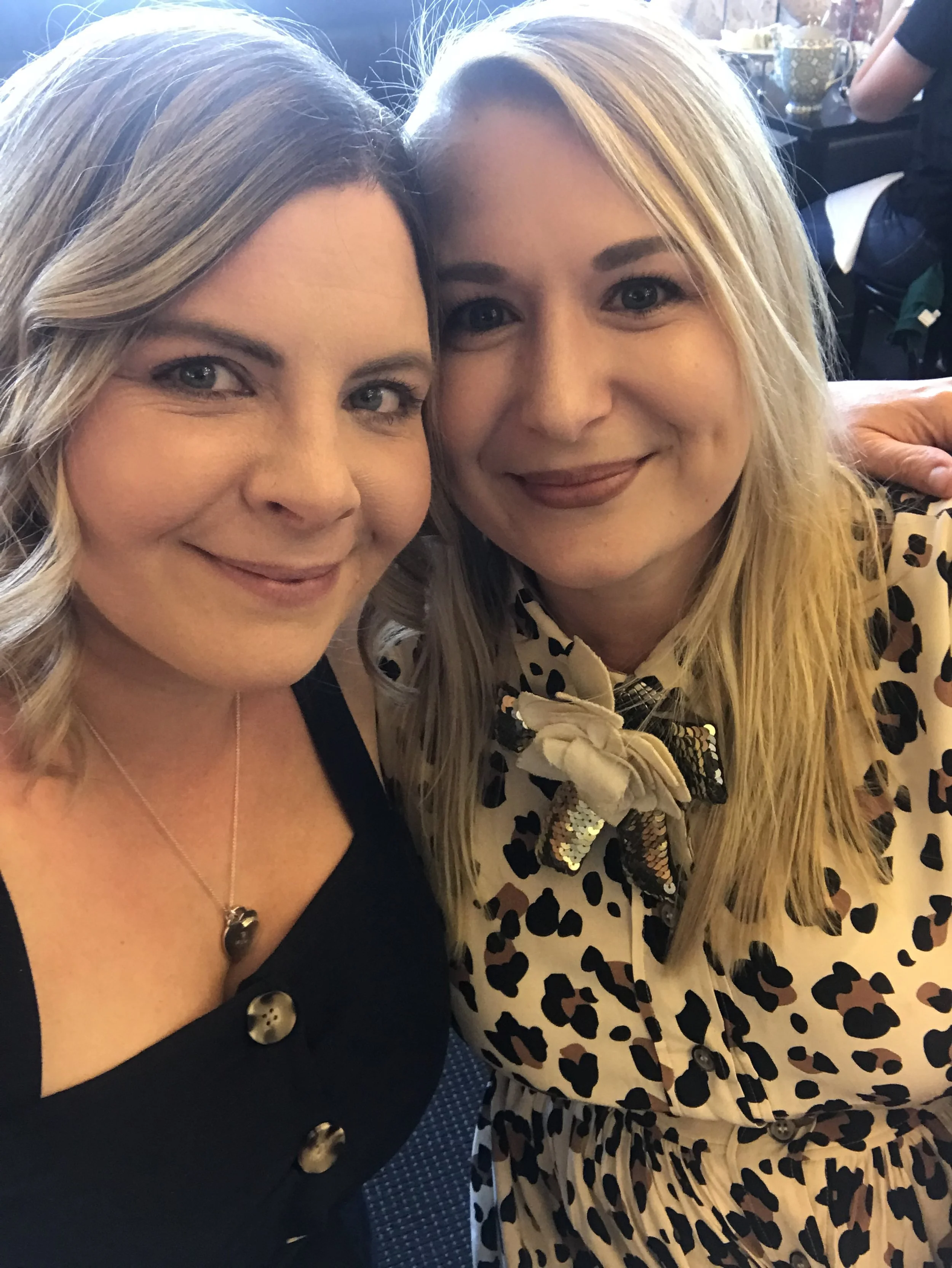 Two women take a selfie, smiling at the camera in a well-lit indoor setting. The woman on the left has blonde hair styled in loose curls and is wearing a black top with buttons and a silver heart necklace. The woman on the right has straight blonde hair and is wearing a cream-colored dress with a leopard print pattern and a bow tie with sequins.