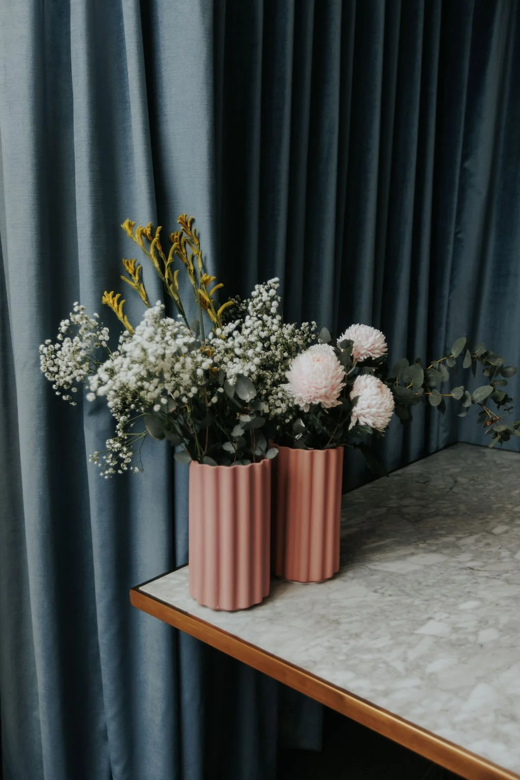 Pink striped vases with white and pink flowers, and green foliage arranged on a marble surface against a blue curtain.