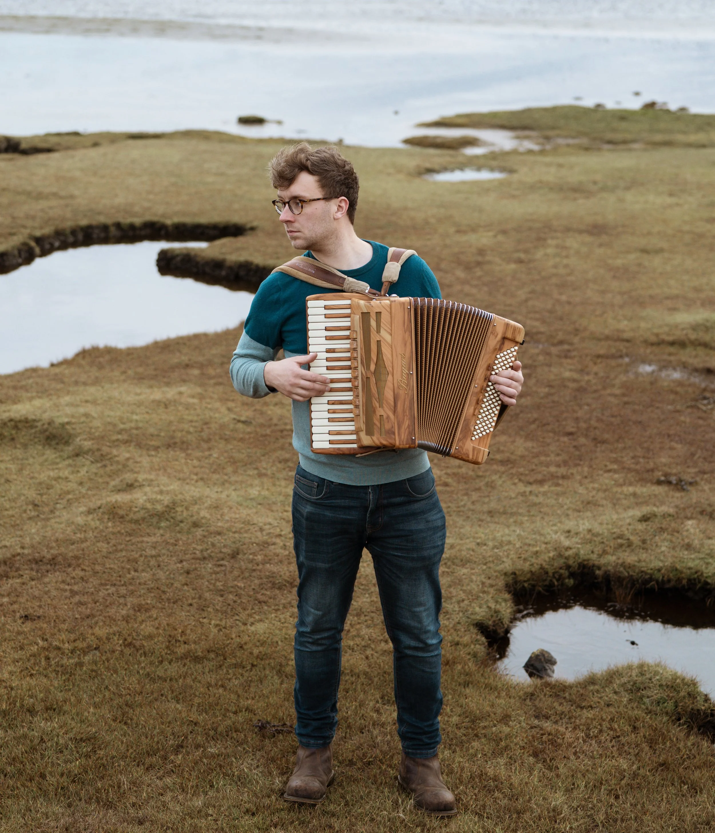A man wearing glasses and casual clothing playing an accordion outdoors on a grassy area near water.