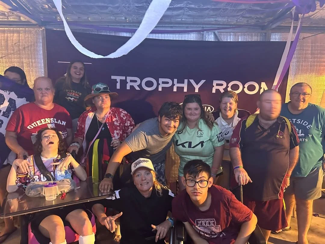 Group of people smiling and posing for a photo inside the Trophy Room, with some holding awards or medals, and a large sign in the background that reads 'Trophy Room'.