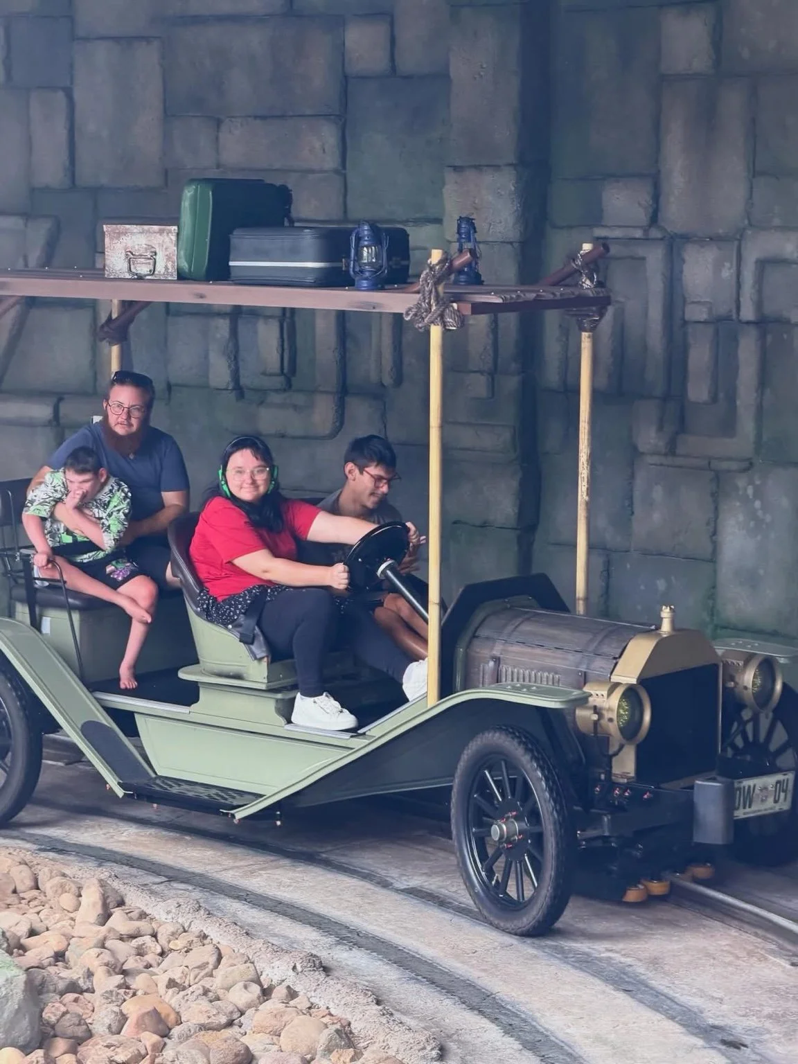 Four people riding an amusement park train with a vintage train design, surrounded by rocky terrain, in front of a stone wall.