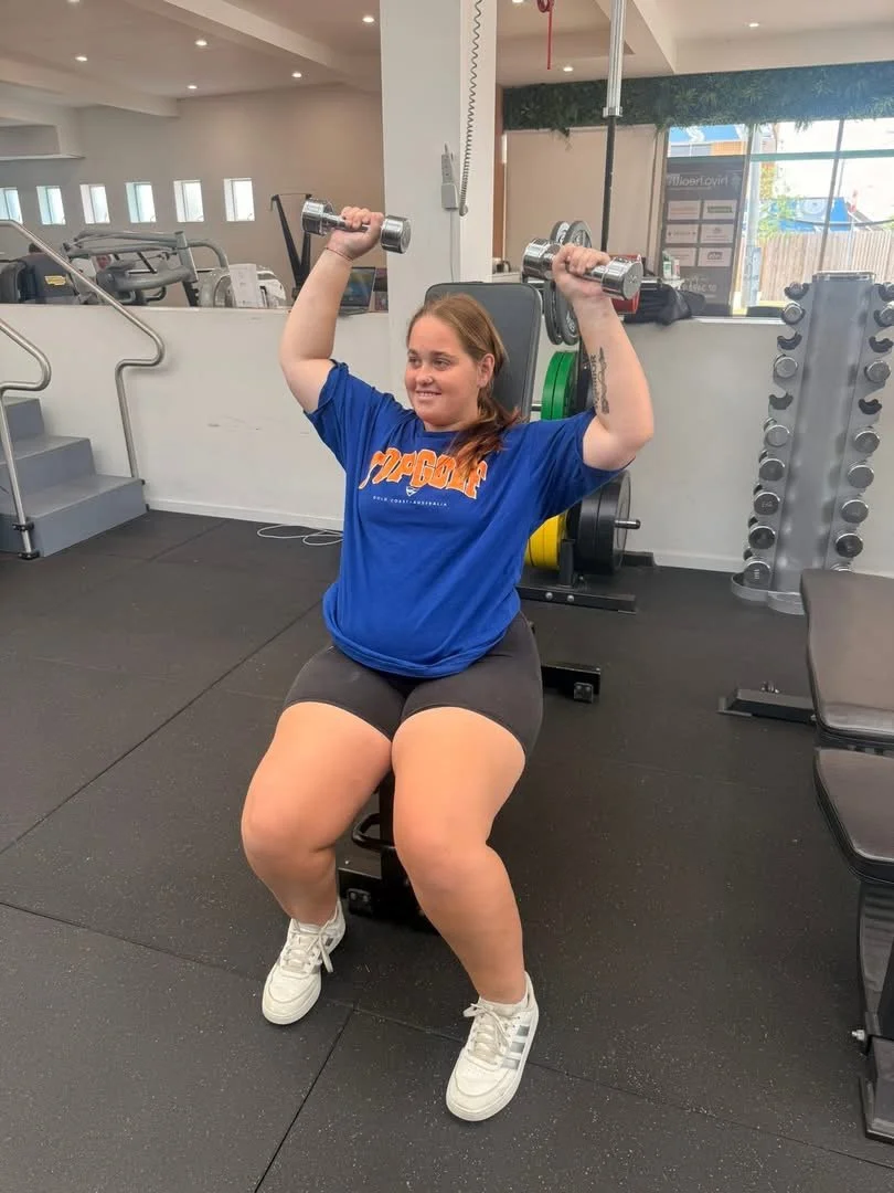 A woman seated on a bench in a gym, lifting dumbbells overhead with both arms, smiling. She is wearing a blue t-shirt, black shorts, and white sneakers, with gym equipment and weights in the background.