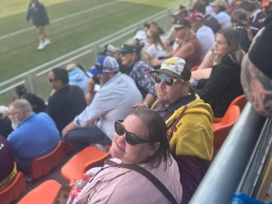 People sitting in stadium seats at an outdoor sporting event. The crowd appears to be enjoying the game, with some wearing sunglasses and hats.