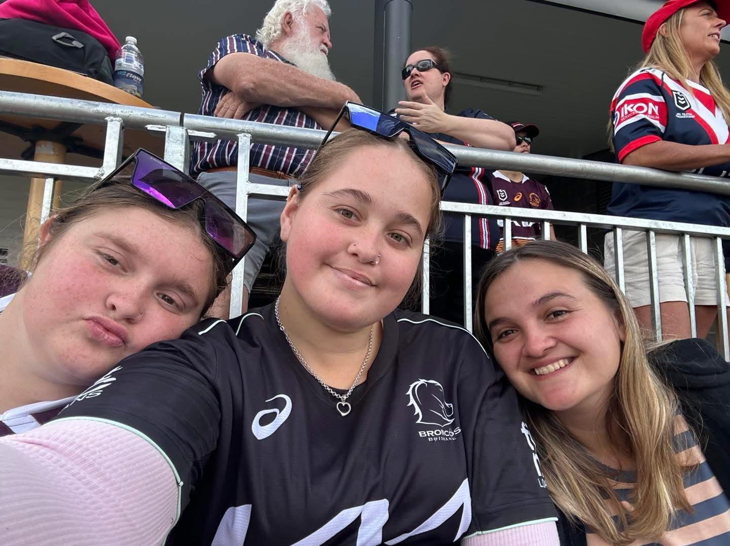 Three young women taking a selfie at a sports event, with a crowd of spectators standing behind a metal railing.