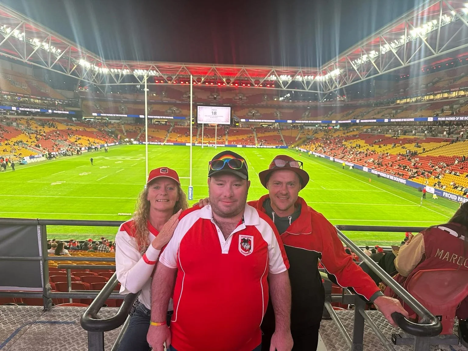 Three sports fans in red and white clothing pose for a photo in a stadium before a rugby game, with a green field and empty seats in the background.