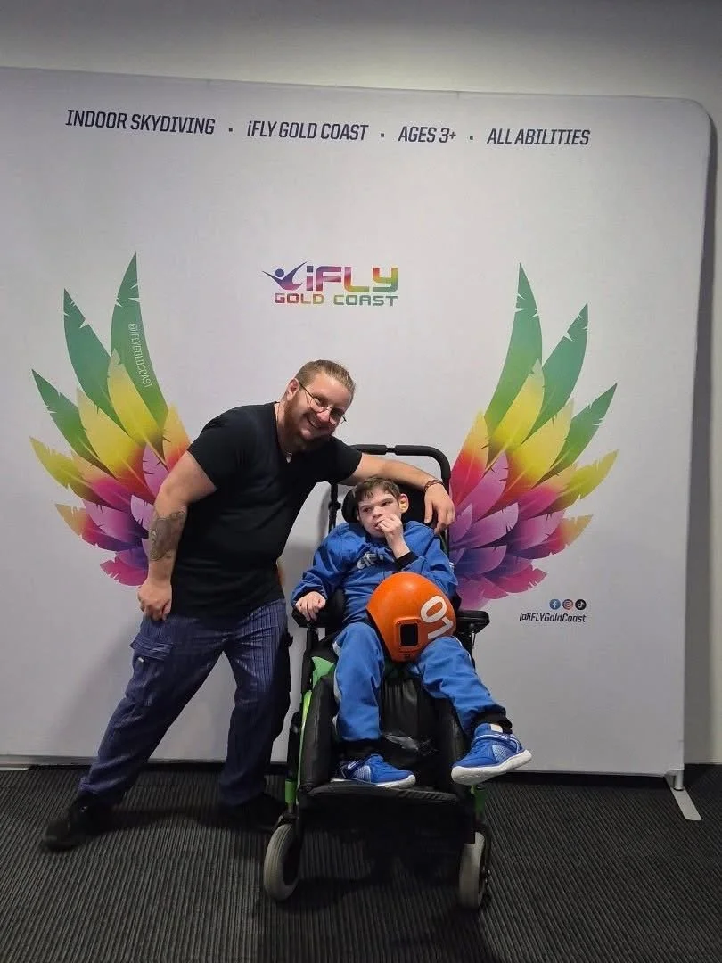 A man and a boy at an indoor skydiving facility with colorful wings on the wall behind them. The boy is sitting in a wheelchair with a helmet, and the man is standing next to him leaning on the wheelchair, both smiling.