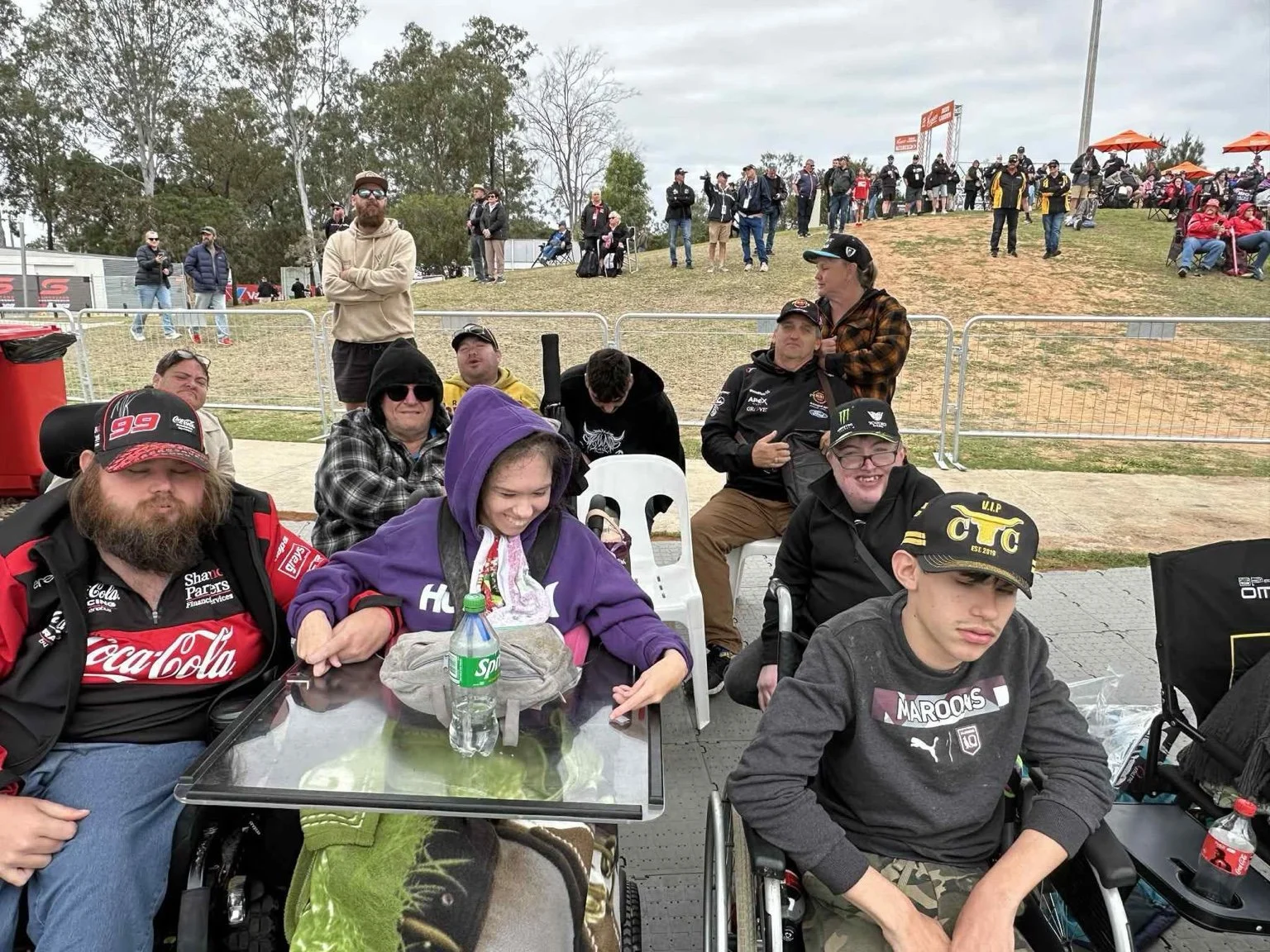 Group of people, including young men with disabilities in wheelchairs, gathered outdoors at a racing event with spectators on a hill in the background.