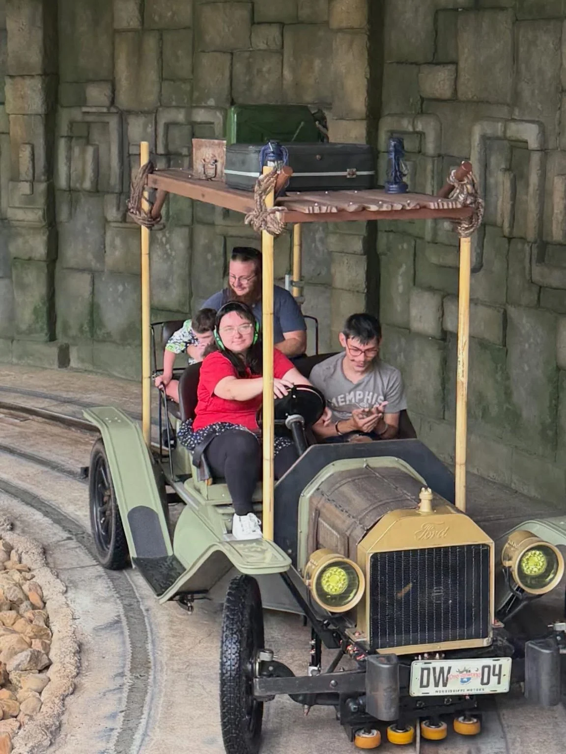 People riding an antique car with a roof structure, inside an indoor amusement park or themed environment built to resemble a stone tunnel or castle wall.