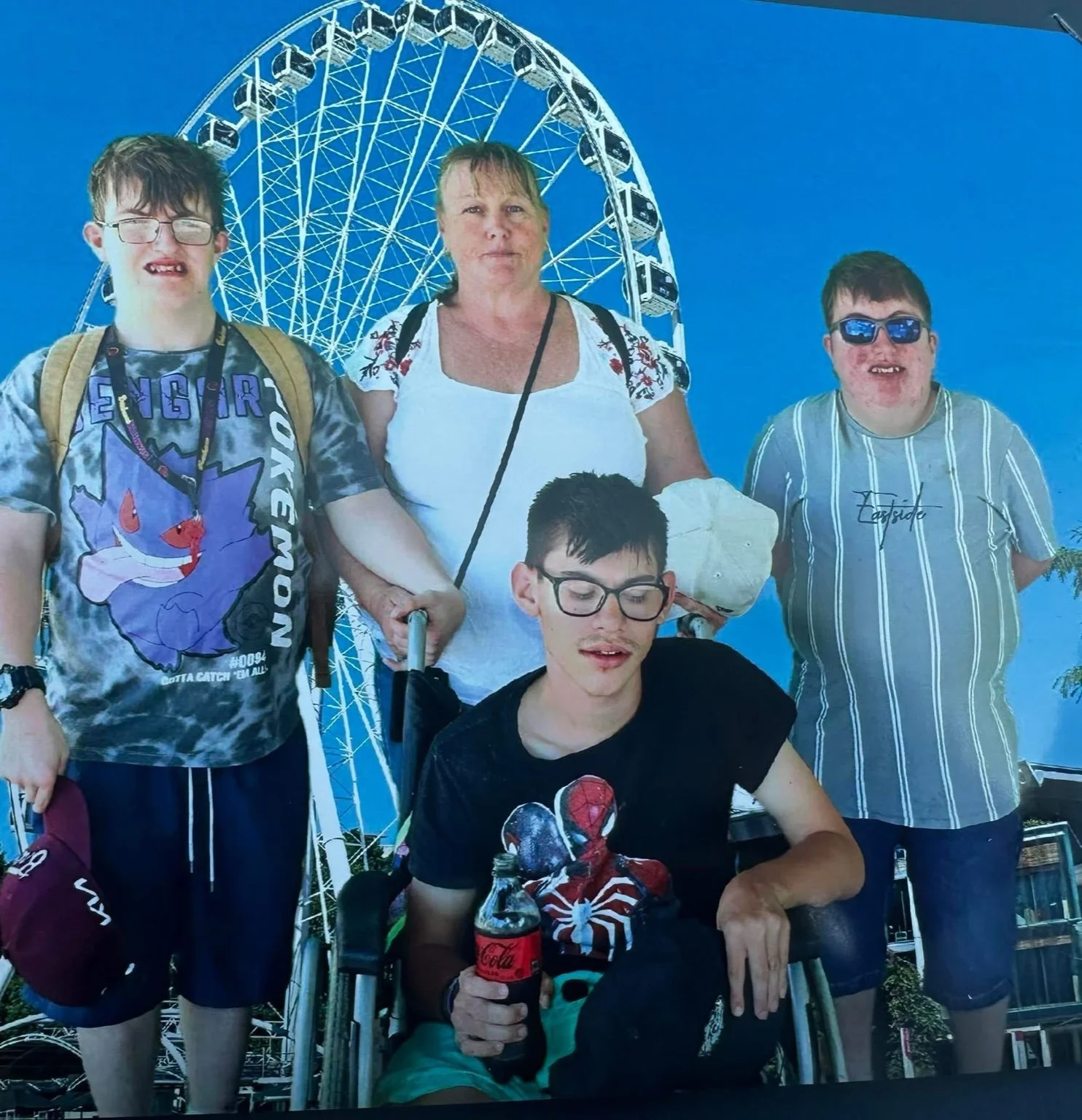 Family group photo at an amusement park with a large Ferris wheel in the background. The group includes a woman and four boys, some with disabilities.