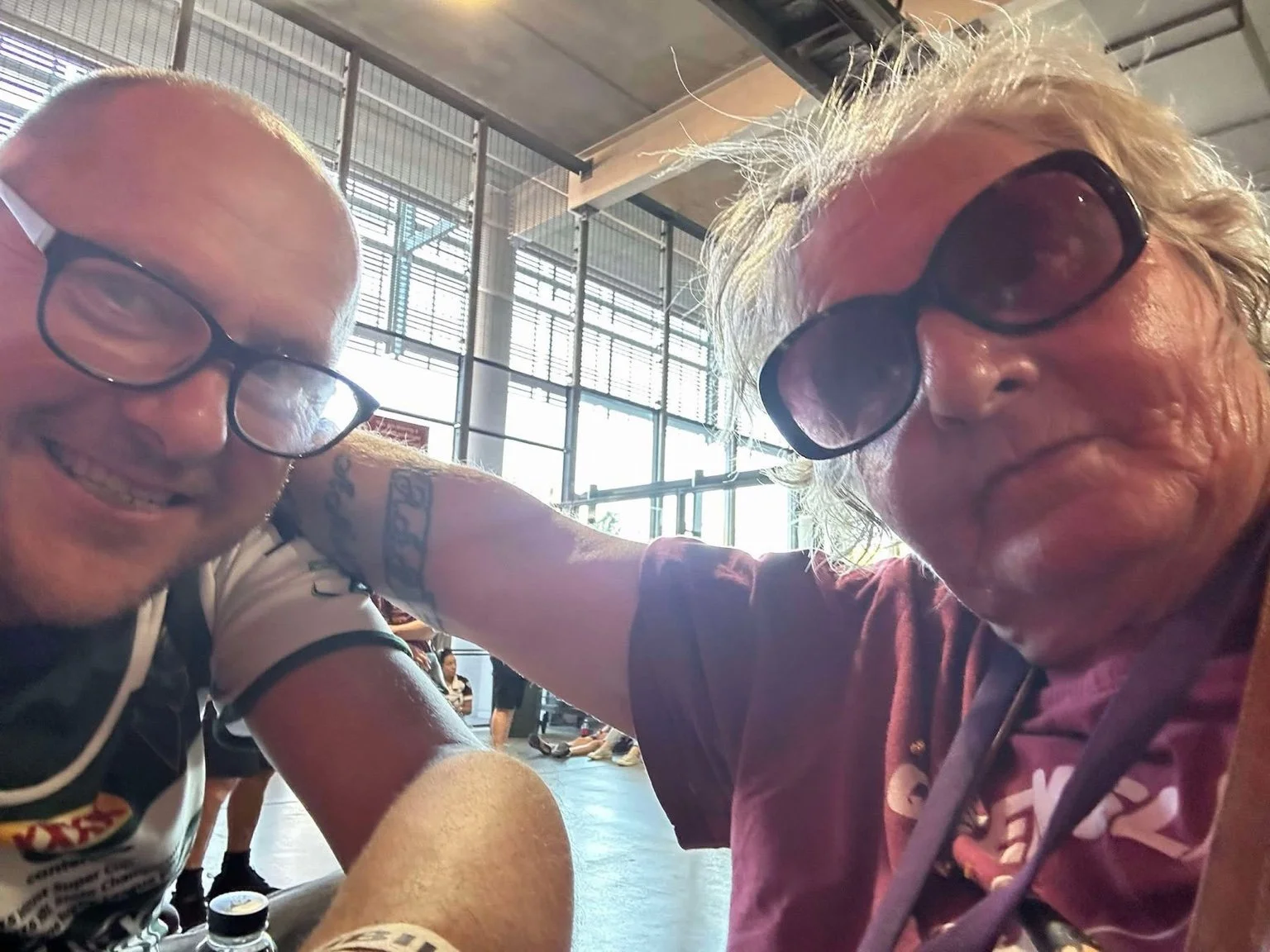 Two men with glasses smiling and sitting close together in a bright indoor setting with large windows; one has blonde messy hair and is wearing a maroon shirt, the other is bald with a tattooed arm.