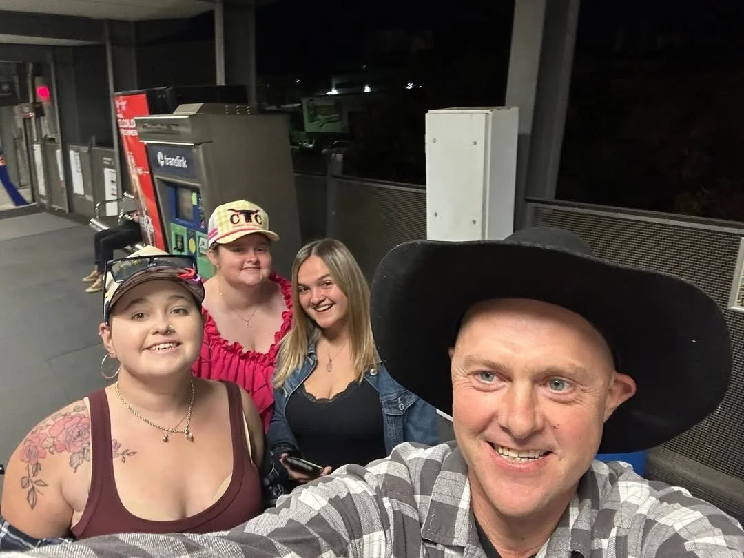 A group of four people taking a selfie at a gas station at night. The group includes a man wearing a black hat and plaid shirt, and three women smiling, one with a floral tattoo, sunglasses, and a cap, and the other two with casual clothing.